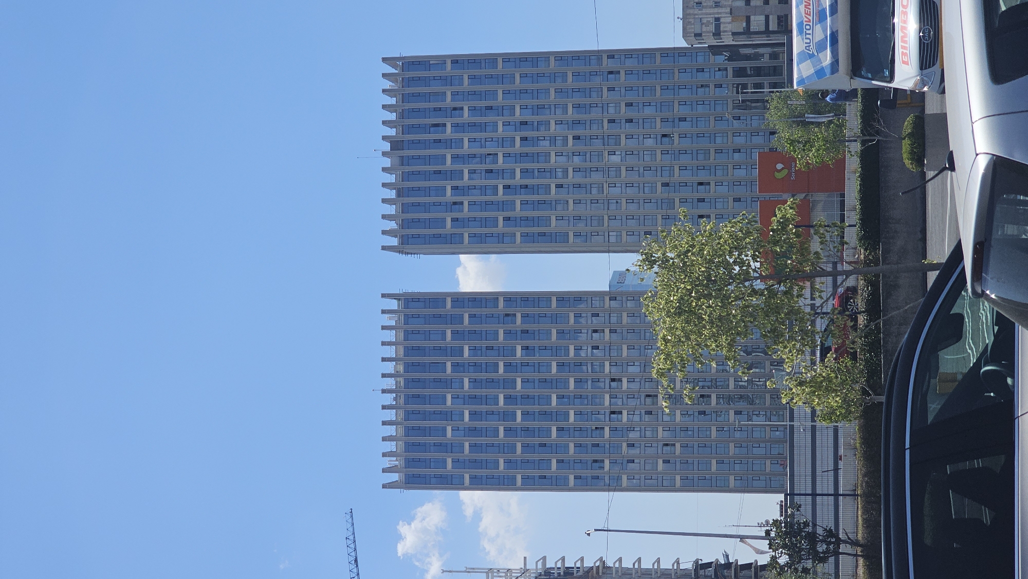 The image displays an urban daytime scene in Puebla, Mexico, under a clear blue sky with scattered white cumulus clouds. Centrally positioned are two identical, modern high-rise towers, characterized by a light-colored structural grid framing numerous rectangular, reflective blue-tinted windows. Each tower culminates in a distinctive, crenelated roofline. To the far left, the metal truss structure of a construction crane is partially visible. In the midground, a substantial tree with light green foliage partially obscures an orange building facade featuring a white "Soriana" logo. Adjacent to this, a white delivery truck, branded with "AUTOVEN" and "BIMBO" logos, is parked or in transit, with a person in a blue shirt discernible next to it. Below these elements, a concrete barrier and a metal fence are present, alongside additional greenery. The immediate foreground is occupied by the partial views of two vehicles: a dark-colored vehicle on the left and a silver vehicle's roof and antenna on the right.