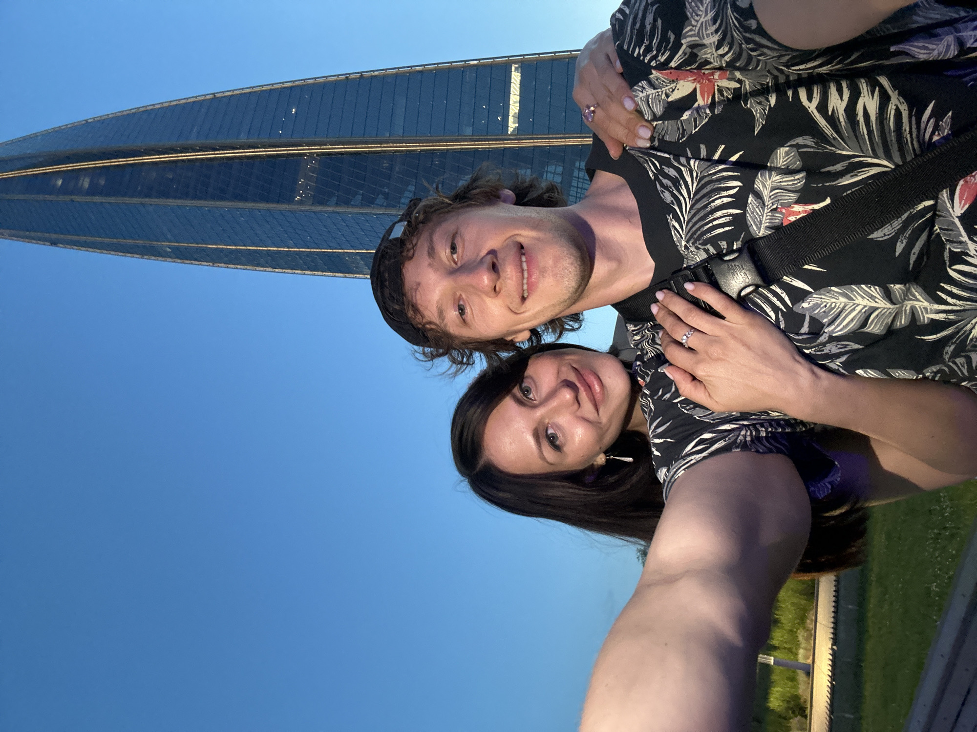 In Saint Petersburg, Russia, a selfie is taken by a couple in front of the Lakhta Center skyscraper. The man, wearing a black baseball cap and a black t-shirt with a tropical print, smiles at the camera. The woman, with dark hair and wearing a black dress, stands to his left, her arm around his shoulders and her hand resting on his chest, showing off her ring. The sky is clear and blue. The Lakhta Center, a modern skyscraper with a glass facade, dominates the background. The foreground shows a grassy area with some lighting fixtures and a bench. The lighting suggests it is evening.