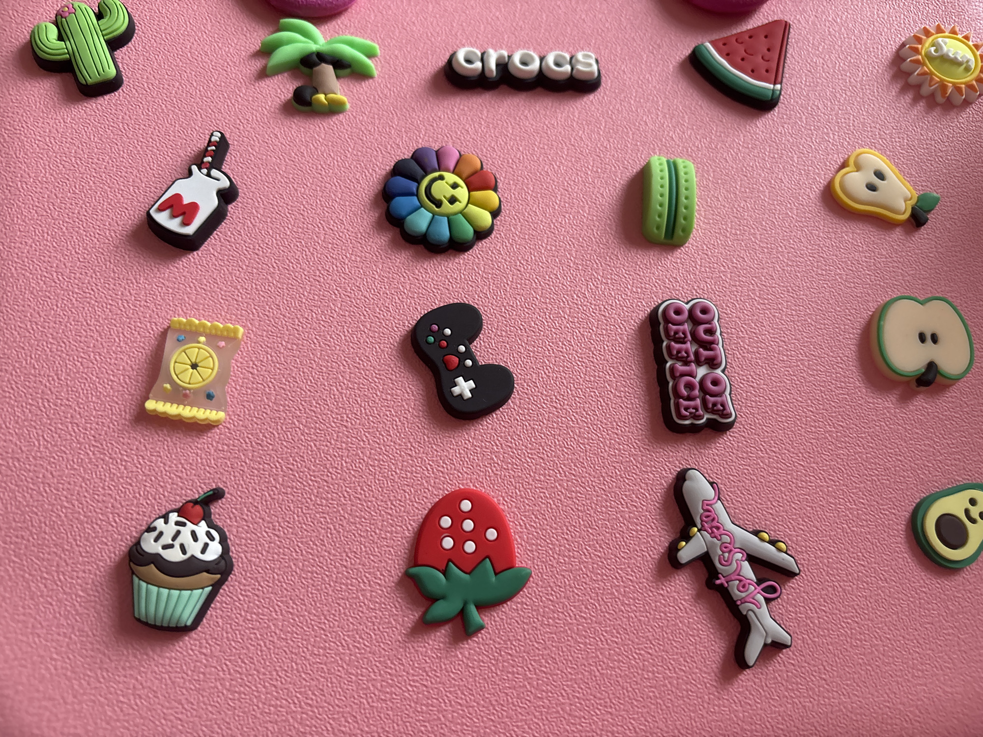 The image shows a collection of colorful, cartoon-like decorative charms arranged on a pink, textured surface. The charms depict various objects and symbols, including a cupcake, a wrapped candy, a cactus, a lollipop, a strawberry, a video game controller, a rainbow flower with a smiley face, the word "crocs" vertically arranged, an airplane with the text "jet setter," a sign that says "out of office," a green macaron, a slice of watermelon, half of an apple, a pear, and a sun-like charm. The charms appear to be made of rubber or PVC material and are positioned at different angles. The background is a solid pink color with a subtle, repeating texture. There is no indication of the city of Georgiyevsk, Russia, or any people or actions in the image.