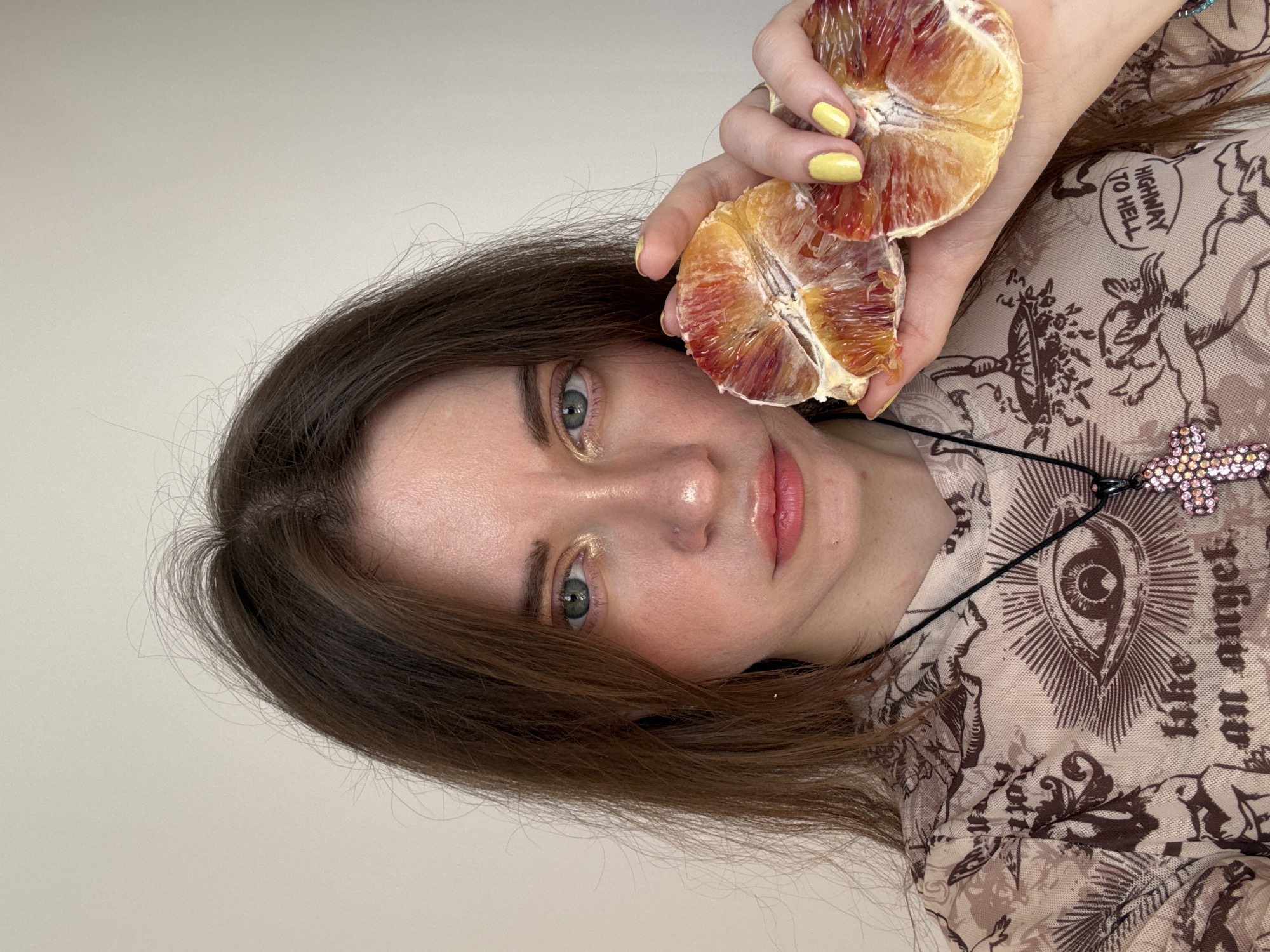 A medium-closeup portrait shows a young woman with light skin, brown hair, and blue eyes. She is holding a blood orange, cut in half, close to her face. Her fingernails are painted yellow. The woman is wearing a light-colored, patterned top with a graphic print and a necklace with a cross pendant. The background is a plain, light-colored wall. There is no information in the image to confirm the location as Moscow, Russia.