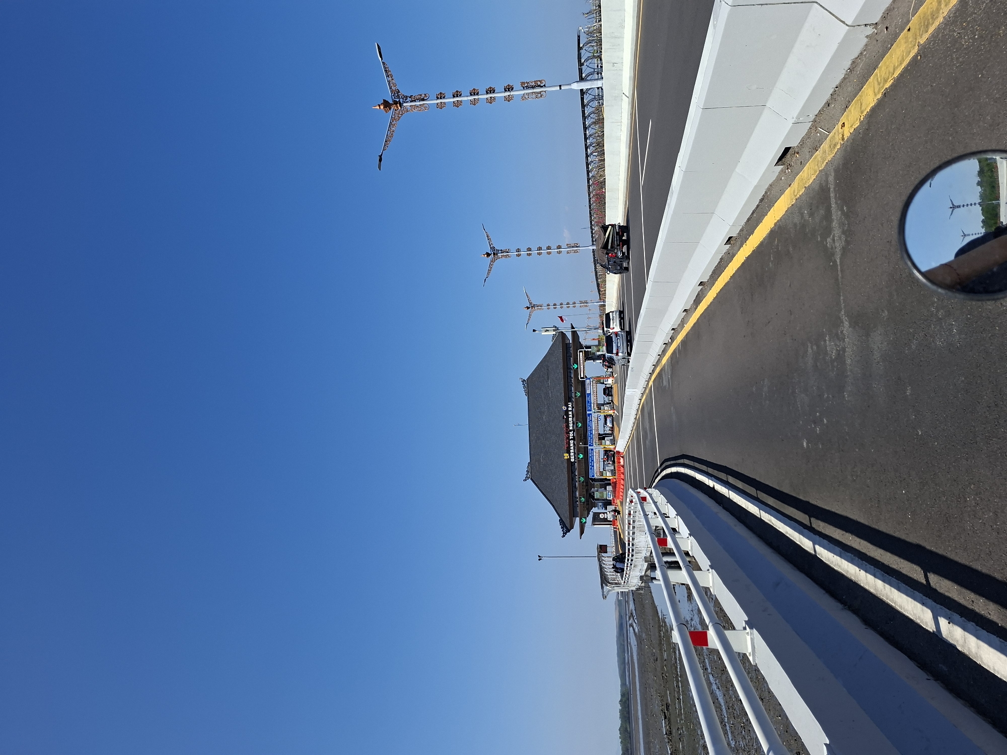 The image shows a toll gate with traditional Indonesian architecture against a clear blue sky. The toll gate is labeled "GERBANG TOL NGURAH RAI" which indicates its location at the Ngurah Rai Toll Gate in South Denpasar, Indonesia. A highway with multiple lanes is visible, with cars and a truck driving on it. On the left side of the highway, there is a body of water with mudflats and a white railing. Several decorative lampposts line the highway, featuring intricate designs and lamps. A reflection of the sky and lampposts can be seen in a motorcycle's side mirror in the foreground. The scene appears to be captured from the perspective of someone riding a motorcycle.