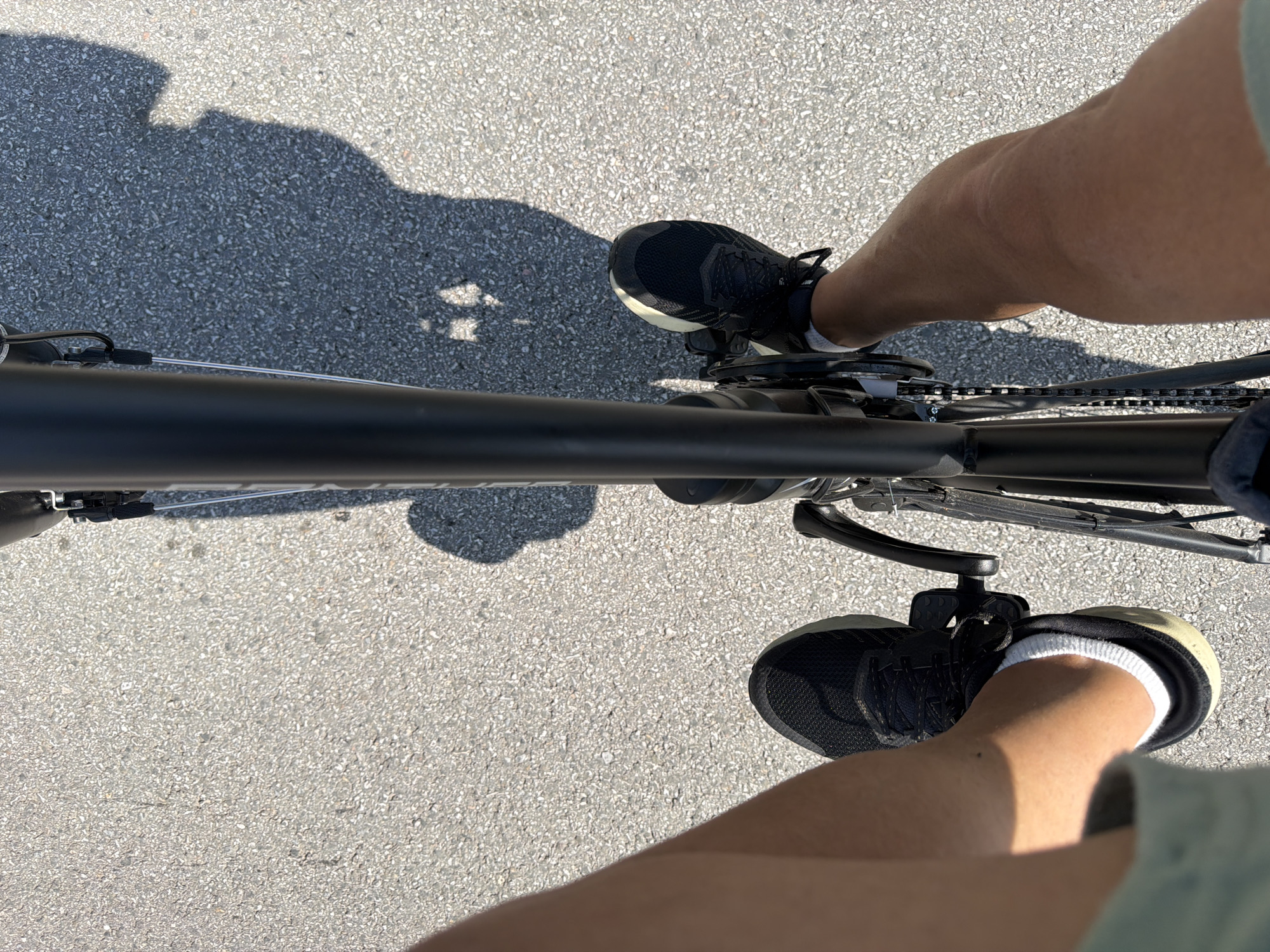 The image shows a first-person perspective of someone riding a bicycle on an asphalt road. The rider's legs and feet, clad in black athletic shoes and white socks, are visible in the foreground, positioned on the pedals of a black bicycle. The shadow of the rider and the bicycle is cast on the road to the right. The environment appears to be outdoors, with a textured asphalt surface. There is no discernible urban or natural landmark to pinpoint a specific location within Kadievo, Bulgaria. The scene captures a moment of cycling activity.