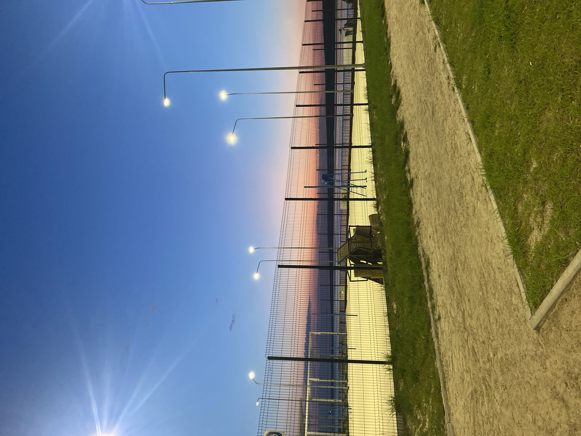 The image depicts a view from Chaikovsky, Russia, possibly near a waterfront or park area. A fence runs along the length of the image, separating a walkway or path from what appears to be a body of water. Streetlights are spaced along the fence line. The sky is a gradient of blue and pink hues, suggesting either sunrise or sunset. A grassy area is present adjacent to the walkway. There are some objects located on the other side of the fence, including what may be chairs and other structures.