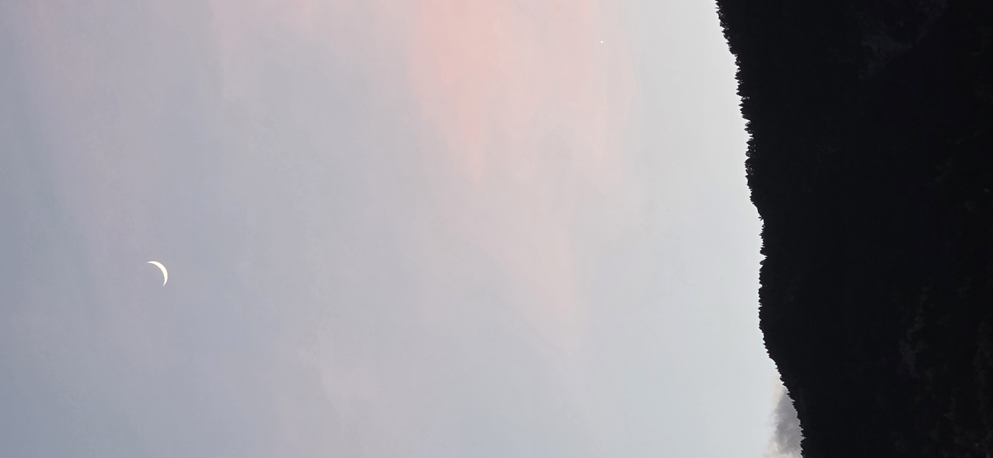The image depicts a sliver of a crescent moon in the sky at twilight. To the right, an out-of-focus, dark, silhouetted mountain looms. Above the mountain, a single star is visible. The sky is a mix of grey and pastel pink hues. Based on the presence of mountains and the description provided, the image likely captures a scene near Benasque, Spain, but no specific landmarks are visible to confirm the location. No people, actions, or interactions are visible in the image.