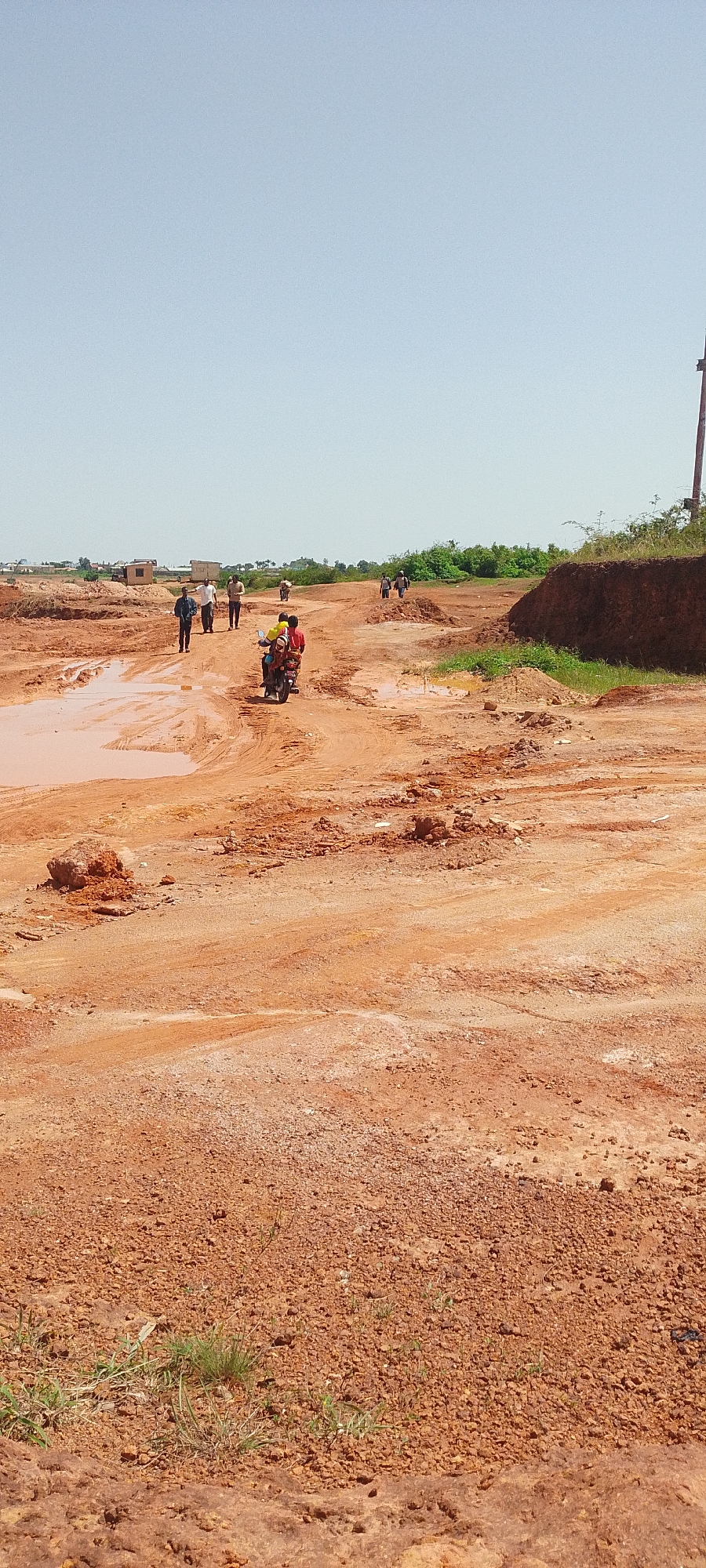 The image depicts a dirt road in Reifild-Ist, Nigeria, characterized by red earth and significant puddles of muddy water, suggesting recent rainfall or poor drainage. Several individuals are present. In the foreground, the terrain is rough and uneven, with loose gravel and clumps of red soil. Further up the road, a group of people are walking in various directions. A motorcycle carrying two individuals, one wearing a yellow shirt and the other a red shirt, is traveling on the road. In the background, more people are walking, and there are scattered structures, possibly buildings, along with green vegetation. The sky is clear and bright blue, indicating a sunny day. The overall scene suggests a rural or developing area undergoing construction or experiencing challenging weather conditions.