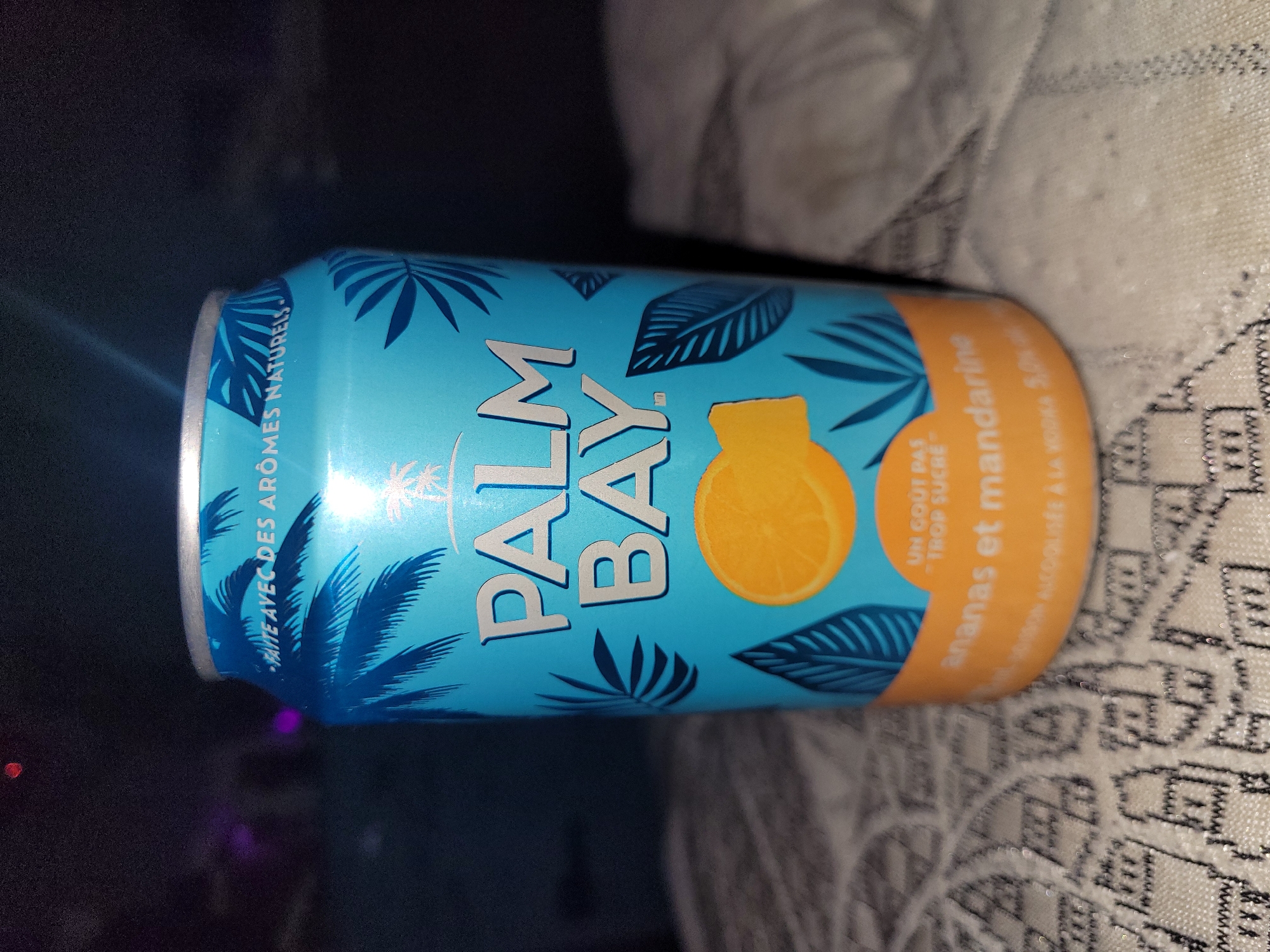 The image shows a can of Palm Bay brand alcoholic beverage, specifically the "ananas et mandarine" (pineapple and tangerine) flavor. The can is light blue with blue palm tree and leaf designs, and an orange section near the top with a graphic of a cut orange or tangerine. The text on the can includes "PALM BAY," "FAITE AVEC DES ARÔMES NATURELS," and "UN GOÛT PAS - TROP SUCRÉ." The can appears to be resting on a surface with a textured, possibly knitted, pattern. The background is a mix of dark and light tones. It is not possible to tell if it's in Calgary, Canada.