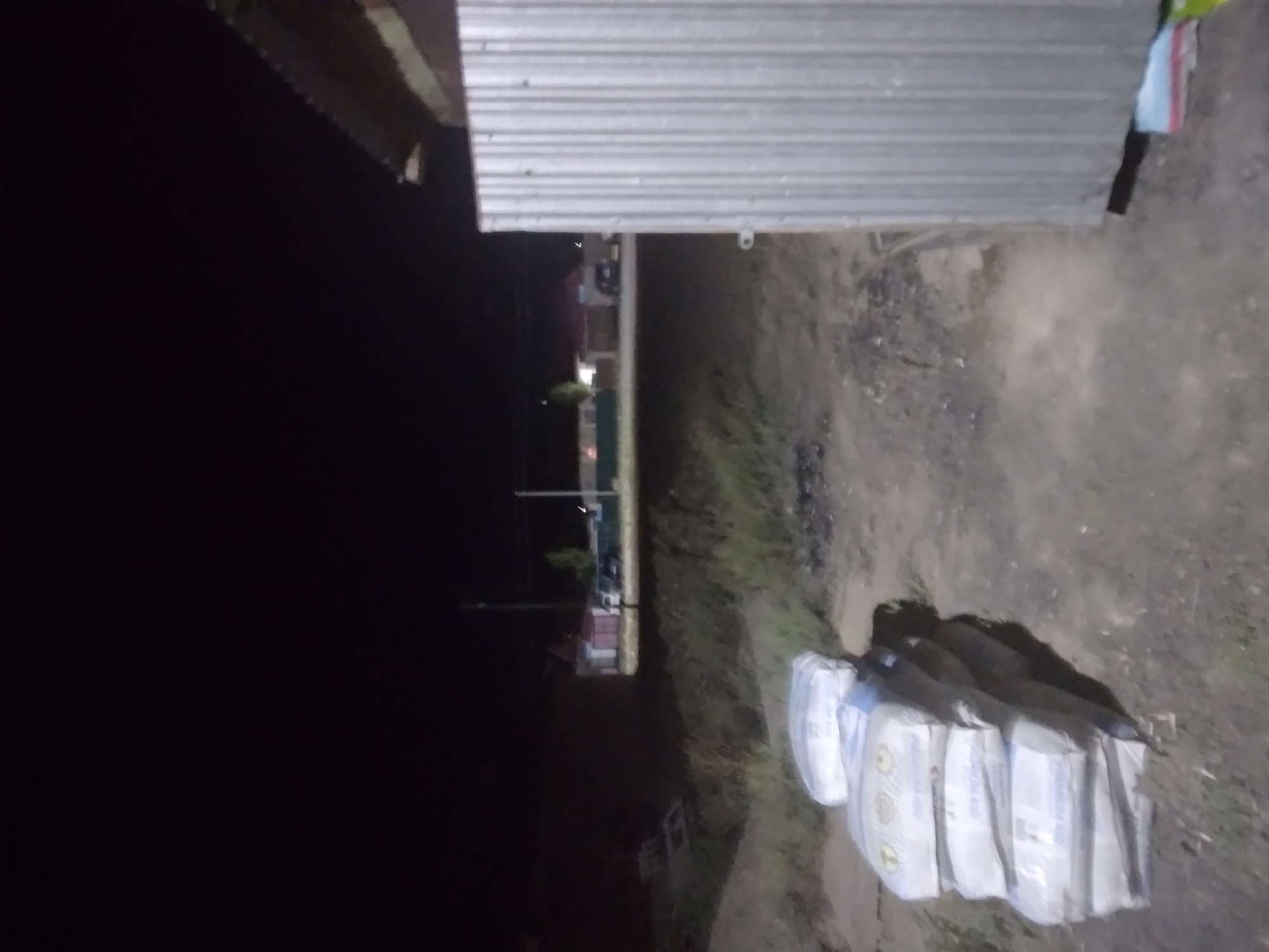 The image, taken at night in Ayagöz, Kazakhstan, shows a stack of eight white cement bags in the foreground. The bags are arranged in two columns, with four bags in each column, and are lying on the ground. Behind the bags, a corrugated metal wall is visible on the right side of the frame. In the background, there are several houses with lights on, and a few vehicles. The overall environment appears to be a rural or semi-urban area at night. The lighting is dim, suggesting artificial light sources are present, possibly from the houses or streetlights.