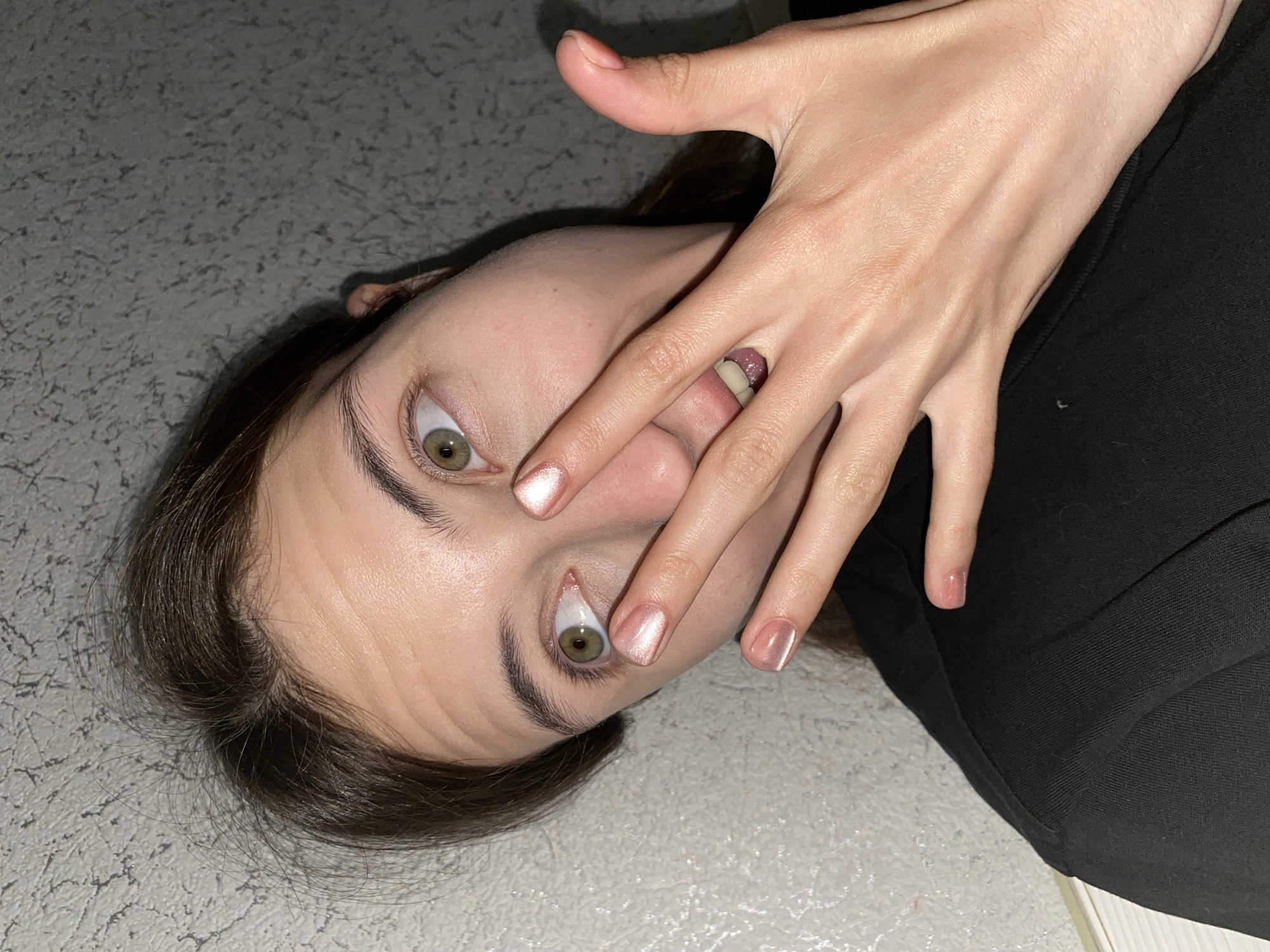 A close-up shot shows a person lying on a textured grey surface. The person's head is tilted, and their face is partially obscured by their hand, which is positioned over their mouth and nose. The visible eye is wide open, with a green iris and dark eyelashes. The person has dark brown hair, and a portion of their ear is visible. Their fingernails are painted with a pearlescent pink polish. The person is wearing a black garment. The lighting suggests an indoor setting, possibly at night, with shadows cast by the hand and head. The overall emotion conveyed is one of surprise or alarm. There is no visible text in the image.