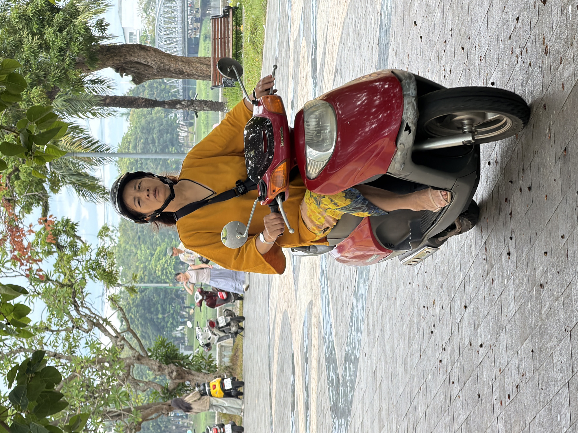 A middle-aged Vietnamese woman wearing a yellow top, patterned pants, and a black helmet is riding a red and silver scooter. She is looking straight ahead with a neutral expression. The scooter has a black windshield and silver handlebars. She is wearing beige sandals. In the background, several other scooters are parked, and two women and a man are walking. The scene takes place on a paved walkway with a pattern of interlocking grey tiles. There are trees and lush greenery surrounding the walkway, and a body of water and a bridge are visible in the distance. The weather appears overcast. The location is Thuận Hóa Ward, Vietnam.