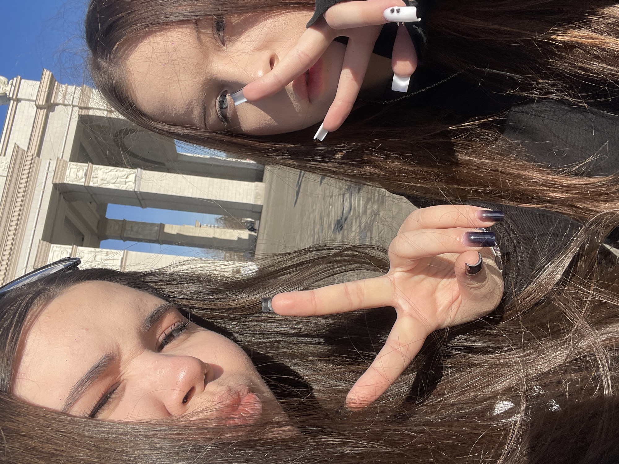 Two young women pose for a selfie. The woman in the upper portion of the frame makes a "peace sign" with long white, ghost-themed acrylic nails. The woman in the lower portion of the frame makes a similar hand gesture with long dark blue acrylic nails. The background reveals a building with classical architecture, with possible stone columns. The image may have been taken near the Khimki Park Culture and Leisure in Khimki, Russia.