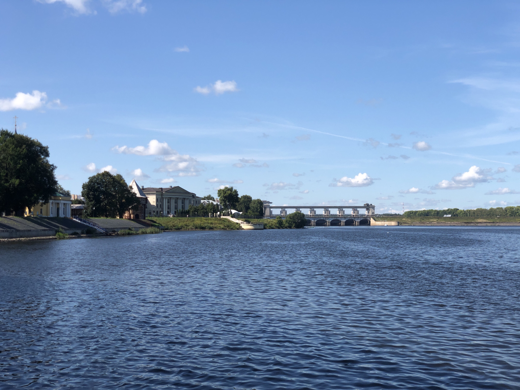 The image captures a serene daytime scene of the city of Fedotovo, Russia, featuring a wide river in the foreground and buildings and a dam in the distance. The river's surface is a deep blue, rippled by a gentle breeze. On the left bank, a row of trees partially obscures a yellow building with white columns and a tall, slender tower topped with a cross. Adjacent to this is a larger, lighter-colored building with classical architectural elements, also bordered by trees. Further down the riverbank, a paved walkway leads towards a large dam structure that spans the width of the river. The dam is made of concrete and features multiple openings and a walkway on top. The horizon is lined with lush green trees and a clear blue sky dotted with scattered white clouds. The overall atmosphere is peaceful and suggests a clear, sunny day. There are no visible people or animals in the scene.