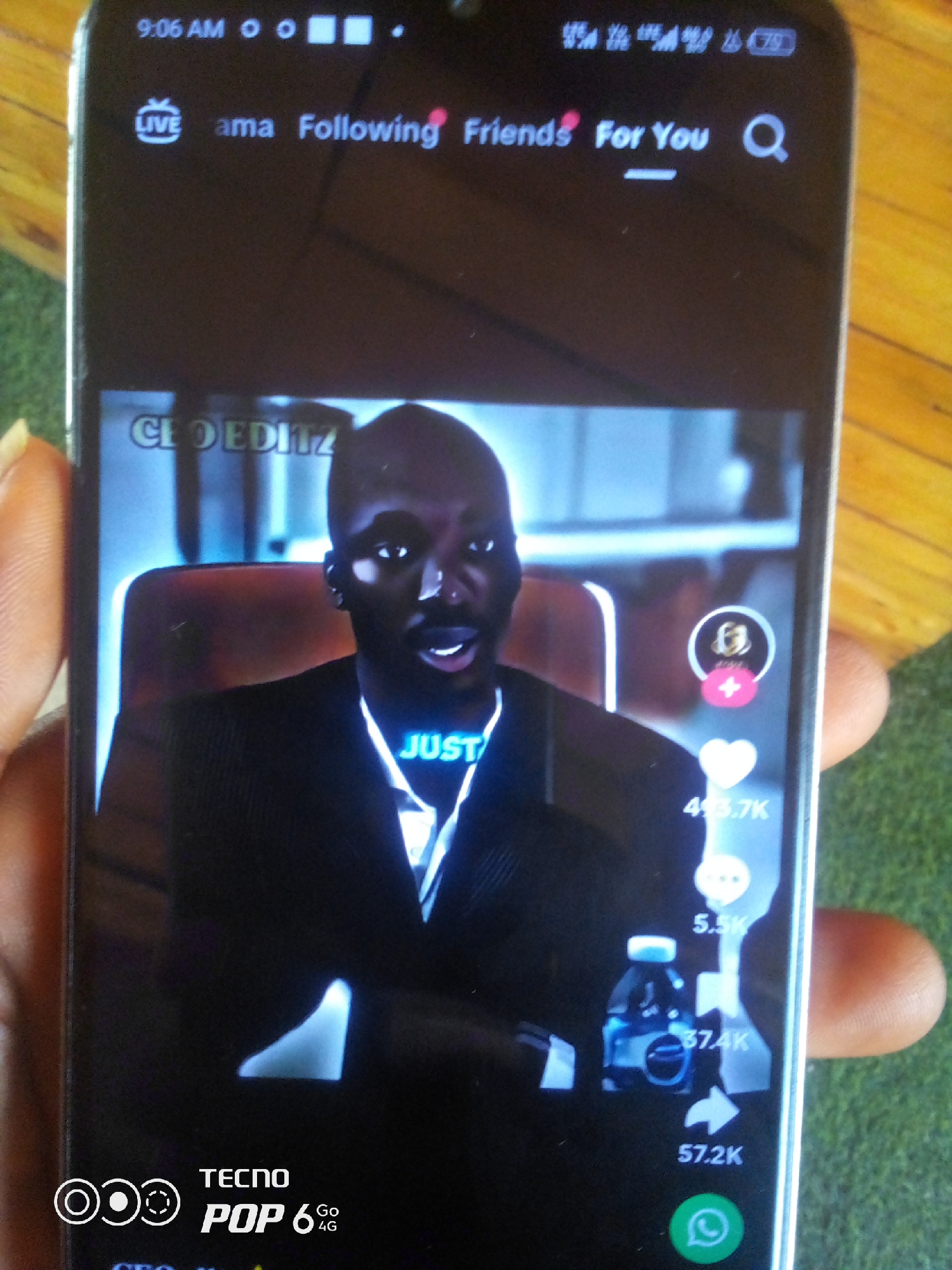 The image is a close-up, vertical shot of a smartphone screen, held by a person's fingers visible on the left. The phone displays a video on a social media platform, likely TikTok, indicated by the user interface elements. The video features a Black man in a suit, with text overlays like "CEOEDITZ" and "JUST". On the right side of the screen, there are interactive icons for likes (a heart with a count of 413.7K), comments (a speech bubble with a count of 5.5K), and shares (an arrow with a count of 57.2K). A bottle of what appears to be a beverage is also visible in the video. The top of the phone screen shows the time "9:06 AM" and network status icons. At the bottom of the screen, the "TECNO POP 6 Go 4G" branding is present. The background outside the phone appears to be a wooden surface and a green, grassy area. The image is taken indoors or outdoors.