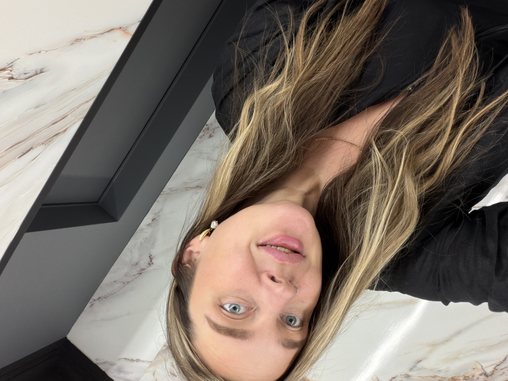 A portrait photograph features a young woman with long, flowing light brown and blonde highlighted hair, depicted in an unusual, rotated orientation. She gazes directly at the camera with bright blue eyes and a subtle, friendly smile, her head tilted slightly. Her complexion is fair, and her lips are a natural light pink. She wears a black top or dress, and a delicate gold chain is visible around her neck. Notable accessories include a silver nose ring and a distinctive earring on her right ear, which combines a pearl drop with a gold-colored leaf accent.
The background comprises a white surface with reddish-brown marble-like veining, contrasted by sharp, dark gray architectural elements that suggest a modern indoor setting. The lighting appears soft and even, highlighting her features. The subject's pose indicates she is posing for the photograph. The image provides no specific time of day or weather cues, but the indoor setting is apparent. This scene takes place in Moscow, Russia. No visible text is present in the image.