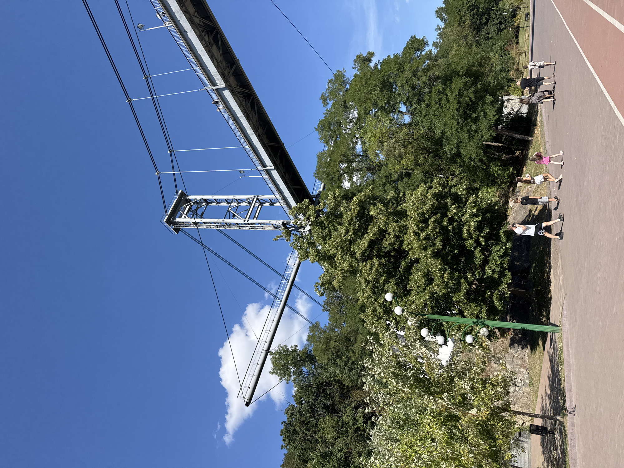 In the city of Unknown, Unknown, on a clear day with a vibrant blue sky and a few scattered clouds, a suspension bridge dominates the upper portion of the image. The bridge's grey metal structure is visible, supported by a tall, angular tower. Suspending cables stretch across the scene, connecting to the bridge's deck. Below the bridge, lush green trees form a dense canopy, obscuring the ground on the far side of a paved pathway.
On this pathway, a group of people are walking. To the left, a person in a white t-shirt and black shorts is walking away from the camera. To their right, a group of children, seemingly a family, are walking together. A taller individual in a black t-shirt and shorts is pushing a stroller or cart with a person in a patterned dress walking beside them. Further to the right, another person in a black and white patterned outfit walks in the same direction. The pathway is a reddish-brown color with a white stripe on the right edge, suggesting it may be a park or recreational area. A tall green lamppost with multiple lights stands to the left of the pathway, amidst the trees. The ground behind the pathway is covered in stones and more greenery.