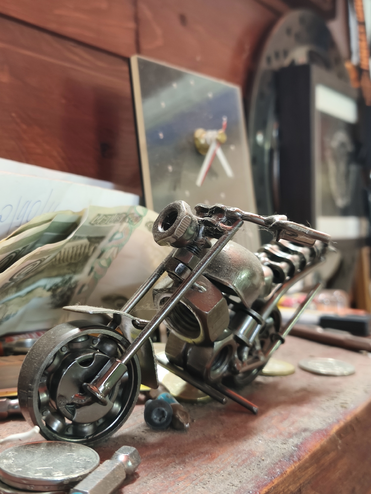 The image depicts a miniature motorcycle sculpture constructed from metal nuts, bolts, and bearings. It is placed on a cluttered wooden surface, possibly a workbench or shelf, with various objects scattered around, including coins, tool bits, and paper currency. In the background, there is a wall with a wooden finish and other items, such as a clock and a framed object, visible in a blurred state. The composition suggests an indoor setting in a workshop or a hobbyist's space. The location is specified as Chaikovsky, Russia.