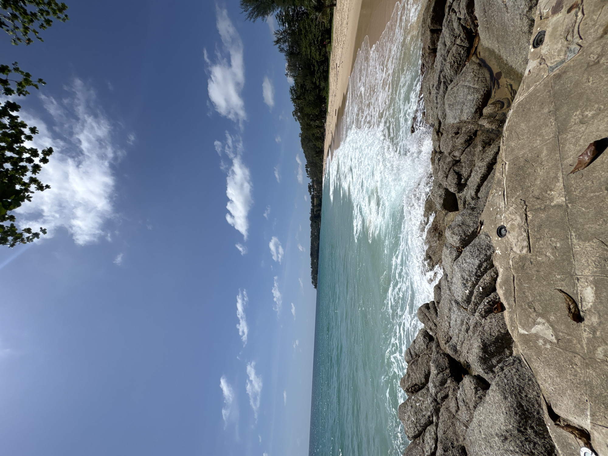 The image depicts a sunny day at a beach in Choeng Thale, Thailand. The foreground is dominated by large, weathered grey rocks. Beyond the rocks, the turquoise water of the ocean meets a sandy beach lined with lush green trees. White waves are breaking on the shore. The sky is a clear blue with scattered white clouds. The scene conveys a peaceful and natural setting.
