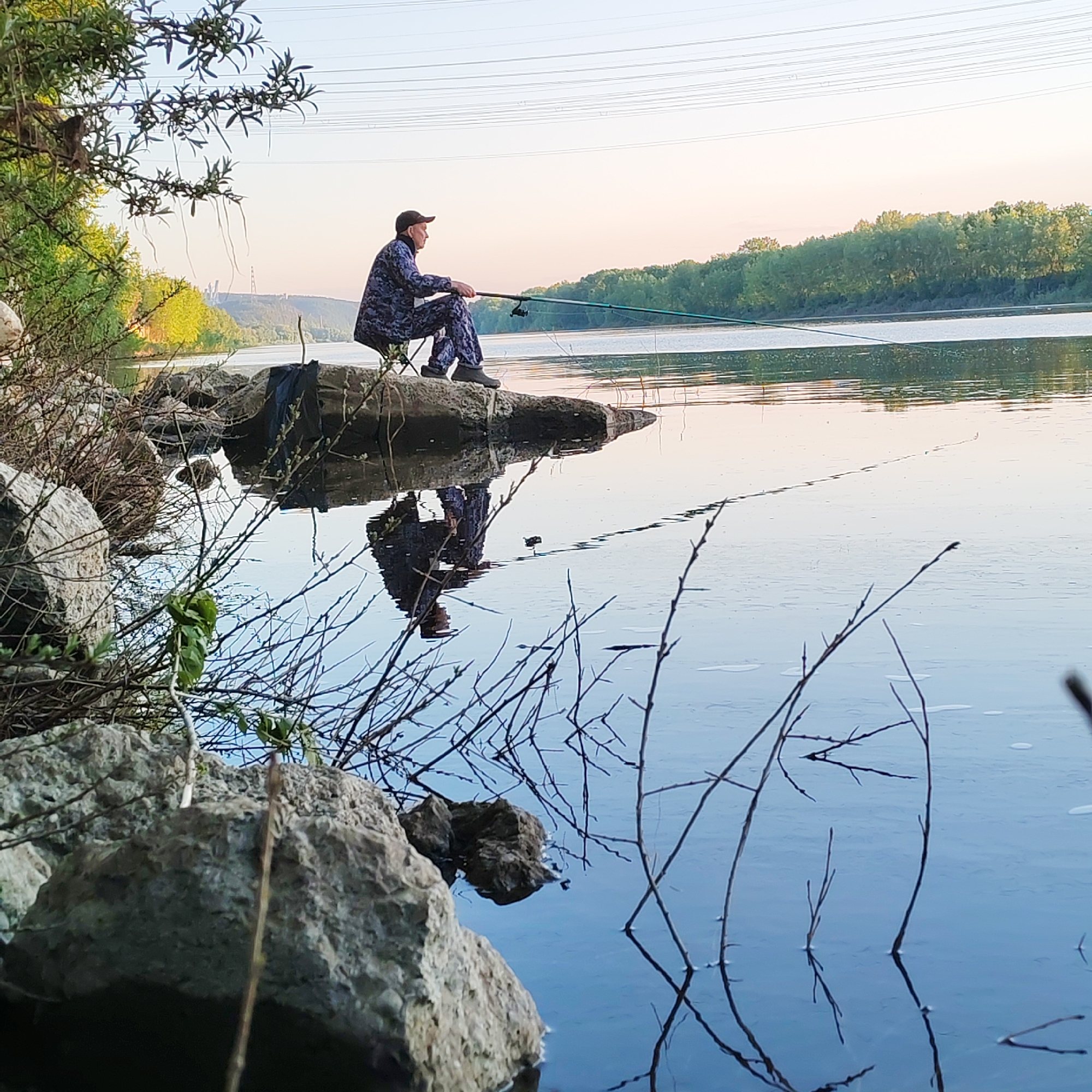The image portrays a serene riverside scene, likely set in Chaikovsky, Russia, featuring a lone individual engaged in fishing. A person, dressed in blue and grey camouflage-patterned clothing and a dark cap, is seated on a small stool or folding chair positioned on a rocky outcrop by the water's edge. They are holding a fishing rod, with the line cast into the calm water, indicating an active fishing session.
The setting appears to be either early morning or late afternoon, characterized by the soft, warm light of the sun low on the horizon, creating a pale, ethereal glow in the sky that transitions from light blue overhead to delicate pink and orange tones. The water is remarkably still, producing a clear, almost perfect reflection of the fisherman and the surrounding riverbanks.
The foreground is dominated by rocks and a tangle of bare branches and new foliage, partially obscuring the view and adding to the natural, untamed feel of the bank. On the distant shore, a dense line of green trees marks the landscape, while further in the background, against the horizon, faint industrial structures or power pylons are visible. Overhead, multiple power lines stretch across the clear sky. A black bag or tarp is discernible among the rocks near the fisherman. The overall mood conveyed is one of peace, patience, and solitude amidst nature. No visible text is present in the image.