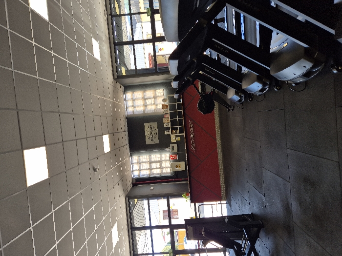 The image displays the interior of a gym, identified as "Instinct Health," located at C. Juliana, 1, 28200 San Lorenzo de El Escorial, Madrid, Spain. The perspective is from a low angle, looking across the main floor towards a reception area and rows of exercise equipment.
The floor is uniformly covered with dark grey or black rubber gym mats arranged in square tiles. To the right, a series of at least four black Technogym treadmills are arranged in a line, facing towards the left side of the frame. A black Concept2 rowing machine is positioned between the treadmills and the central counter. On the far left, a portion of a black leg press machine or similar strength equipment is visible.
In the mid-ground, a prominent reception or service counter stretches horizontally across the scene. The counter's front surface is red with a dark geometric pattern, bearing the white stylized text "INSTINCT HEALTH." Behind the counter, a dark wall features a white rectangular sign with black lettering. Below this sign, multiple open cubby-style shelves contain various small items, including cups, containers, and possibly packaged goods. A small green plant sits on the counter to the left.
The facility is illuminated by both artificial overhead lighting and natural light. The ceiling consists of a grid of square acoustic panels interspersed with numerous square, flush-mounted LED light fixtures. Large windows line the walls, allowing daylight into the space. The windows behind the counter are partially obscured by a patterned translucent film or blinds, while those on the left offer a view of external buildings and outdoor conditions. No individuals are present within the depicted scene.
