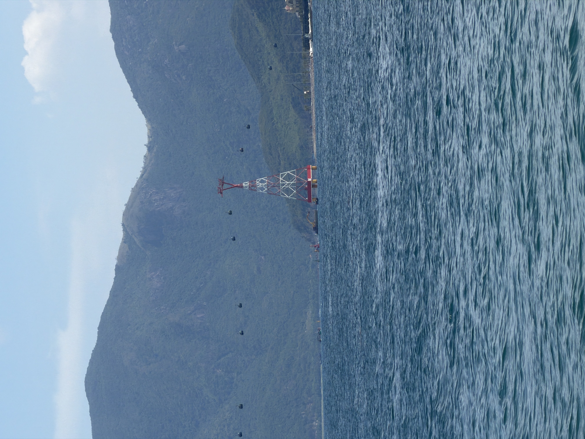 The image depicts a coastal scene. On the left, a mountain range covered in dense greenery rises towards a sky with a patch of white cloud. In the center, a transmission tower with a red and white lattice structure stands near the coastline. Cables for gondolas stretch from the mountainside to the tower, with several gondolas visible along the cables. To the right, the ocean stretches out, its surface reflecting sunlight. In the distance along the shoreline, evidence of a developed city or town can be seen, with buildings and potentially roads visible. This could be Nha Trang Ward, Vietnam, given the image description. The elements of natural landscape and man-made structures, along with the presence of the ocean, suggest a coastal environment with tourism or transportation infrastructure.