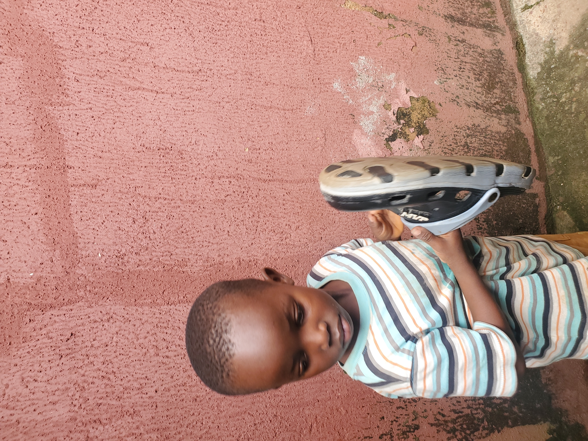 A young boy with dark skin and short hair is lying on his back on a rough, reddish-pink surface. He is wearing a long-sleeved shirt with horizontal stripes in various colors, including blue, orange, black, and white. His eyes are partially closed, and he appears relaxed or possibly sleepy. In his right hand, he is holding a light-colored object with black accents, which looks like a bicycle helmet. The helmet has vents and what appears to be a strap. The background is a textured wall, with some patches of peeling paint and green moss or algae in some areas, suggesting an outdoor setting. The lighting indicates it is daytime. The overall impression is of a casual moment outdoors, perhaps after playing. The location is identified as Jos, Nigeria. No other visible text is present in the image.