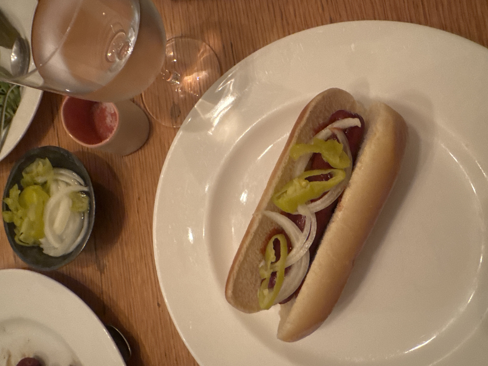 A medium shot of a partially eaten hot dog sitting on a white plate on top of a wooden table. The hot dog is in a bun and topped with sliced onions and green peppers. To the left of the plate are two additional plates and a small bowl containing more sliced onions and green peppers. A glass of liquid is positioned in the upper left corner. The shot was likely taken in New York City, United States of America.