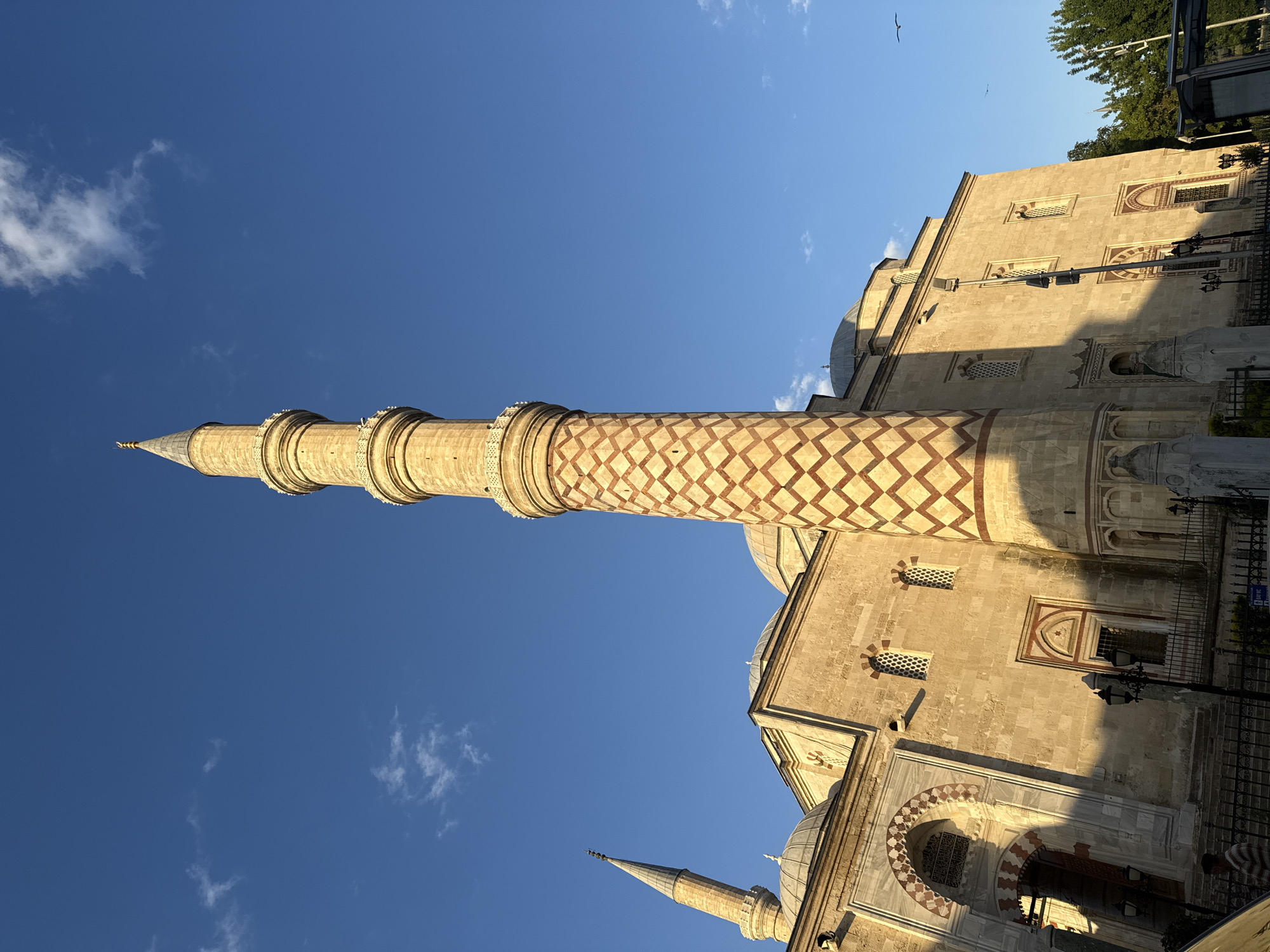 An exterior, low-angle, wide shot captures a historic mosque on a clear, sunny day. The mosque is constructed of light-colored stone and features a prominent, tall minaret with a decorative diamond pattern in red and gold. Another, shorter minaret is visible to the left. The mosque's main structure has several domes, arched windows, and decorative stonework around doorways. A few small clouds dot the bright blue sky. In the foreground, a dark metal fence encloses the mosque's grounds. A few trees are visible to the right, and a portion of a bus shelter is also present. A single person, wearing a striped shirt, walks near the entrance of the mosque. Two birds are flying in the sky. The lighting suggests it is late afternoon, with long shadows cast across the building.