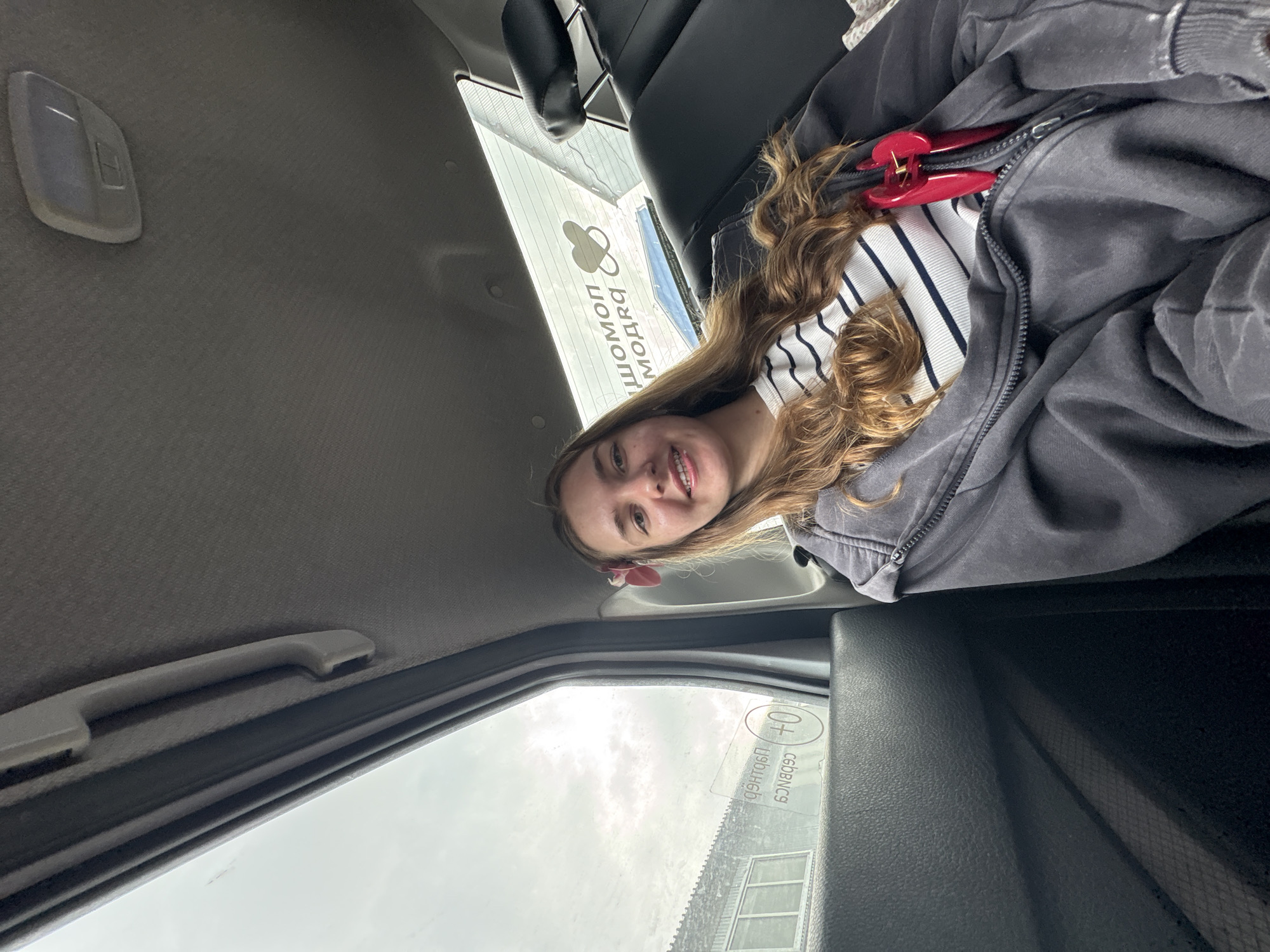 A young woman with long, wavy brown hair is looking at the camera with a smile. She is wearing a striped shirt, a dark gray hoodie, and has a red hair clip in her hair. She is sitting inside a car. The car's interior, including the ceiling, handle, and front window, are visible. Through the front window, a building is seen with the text "ШОПОМ МОДЯ" and "ЦЕНТРБЕ" in Russian, with the city likely being Perm, Russia. The sky appears overcast.