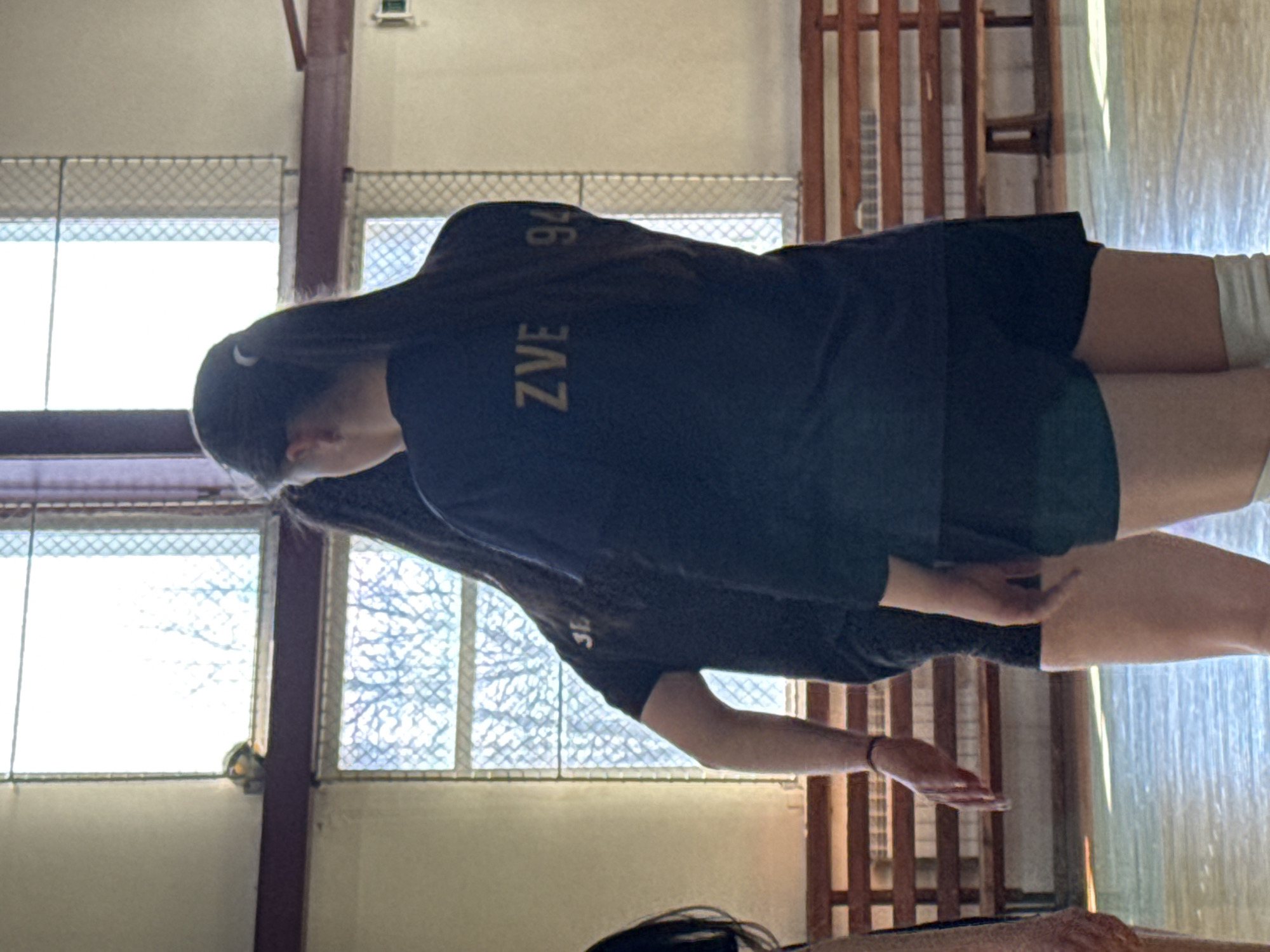 The image depicts a woman in a volleyball uniform with the word "ZVEZ" and the number "94" on the back. She is wearing shorts and knee pads, suggesting she is practicing or playing volleyball indoors, most likely in a gymnasium. The background includes windows with a wire mesh and what appears to be a wooden bench or barrier. The location is unknown but potentially in the city of Sofia, Bulgaria. The woman seems to be in motion, perhaps performing a volleyball skill or drill.