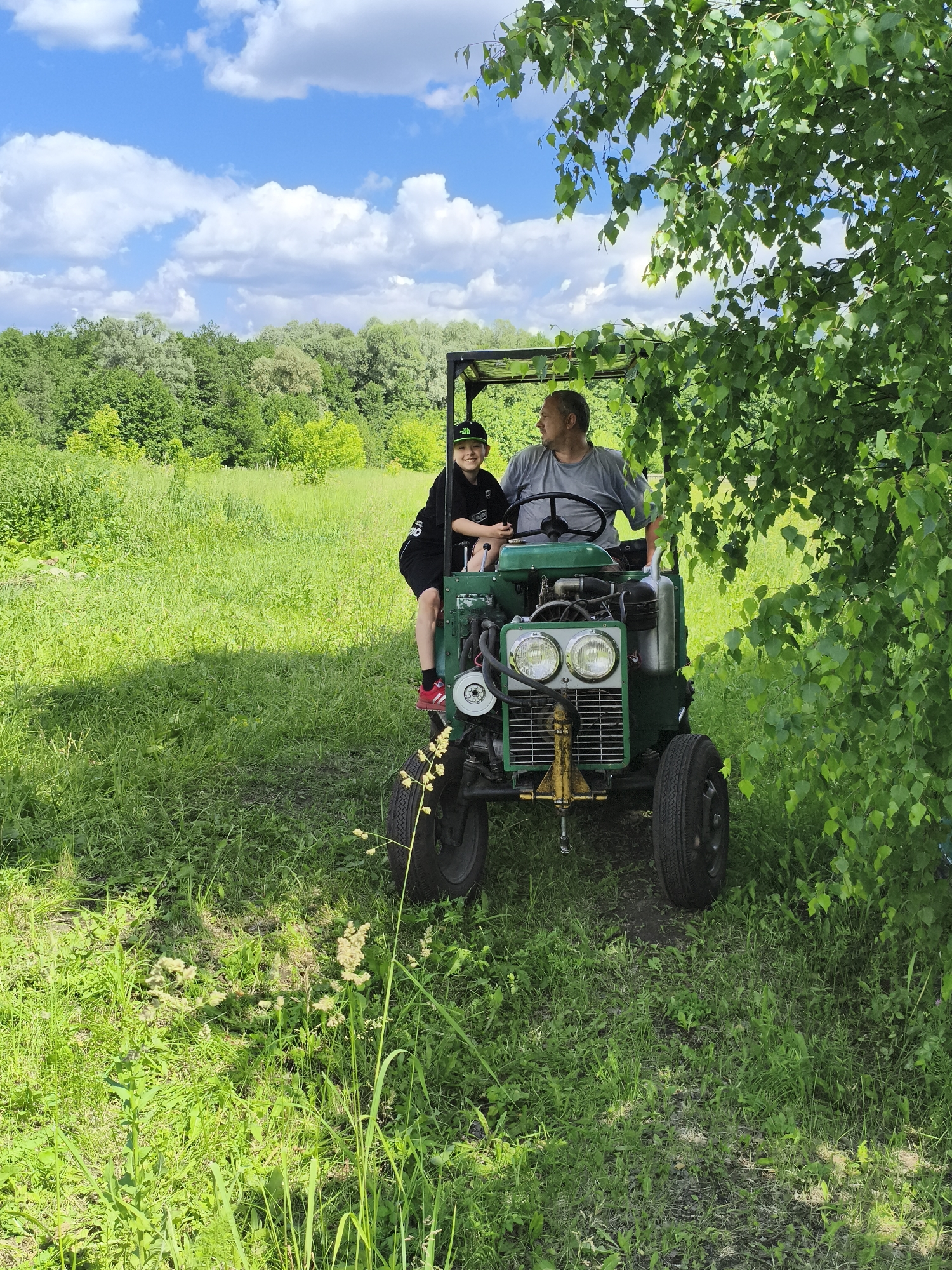 In a grassy field under a bright blue sky with scattered clouds, two individuals are seated on a green tractor. To the right, a mature man with short, graying hair and a grey t-shirt is at the steering wheel. To his left and slightly in front, a young boy wearing a black cap, black hoodie, and black shorts with red and white sneakers is looking towards the camera with a smile. The tractor has two headlights, a yellow tow hitch, and large black tires. The background is filled with lush green trees and vegetation. The location is identified as Ilmino, Russia.