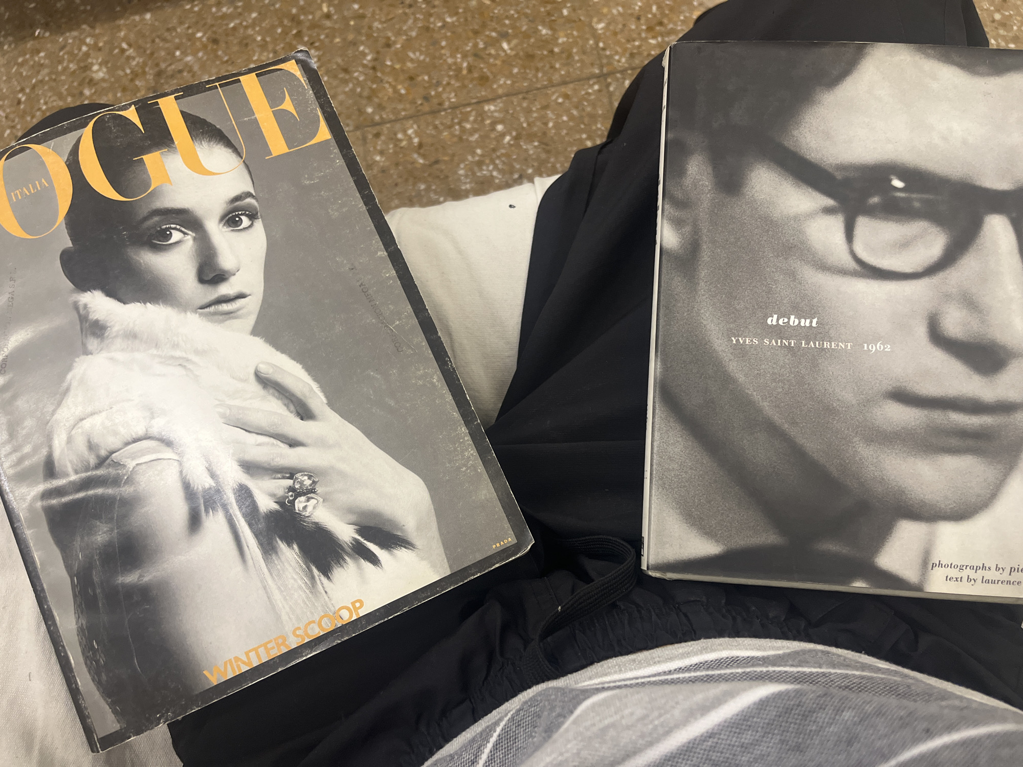 The image shows two books resting on someone's lap. The book on the left is a vintage Vogue Italia magazine with a cover featuring a black and white portrait of a woman in a fur coat. The word "VOGUE" is prominently displayed in yellow lettering at the top, with "ITALIA" beneath. The words "WINTER SCOOP" are written at the bottom. The magazine also has the word "PRADA" on the side. The book on the right is also black and white, featuring a close-up of Yves Saint Laurent wearing glasses. The words "debut" and "YVES SAINT LAURENT 1962" are visible. The bottom corner reads "photographs by pie" and "text by laurence". The person's clothing consists of black shorts with an elastic drawstring and a striped t-shirt. The background is a tiled floor, which is irrelevant to the context of this photo. There is no information to suggest that this photo was taken in Buenos Aires, Argentina.