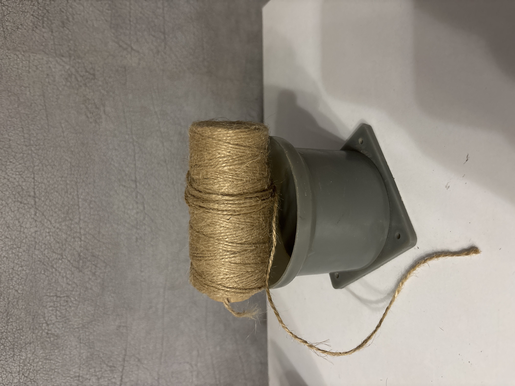 A spool of brown twine rests atop a gray plastic cylinder, which is itself mounted on a square gray base. A length of twine trails from the spool and drapes down the side of the cylinder onto a white surface. The background is a textured gray wall. The scene appears to be indoors, on a white table or shelf. The twine is approximately 3 inches in diameter. The cylinder is approximately 4 inches tall, and the base is approximately 6 inches square.