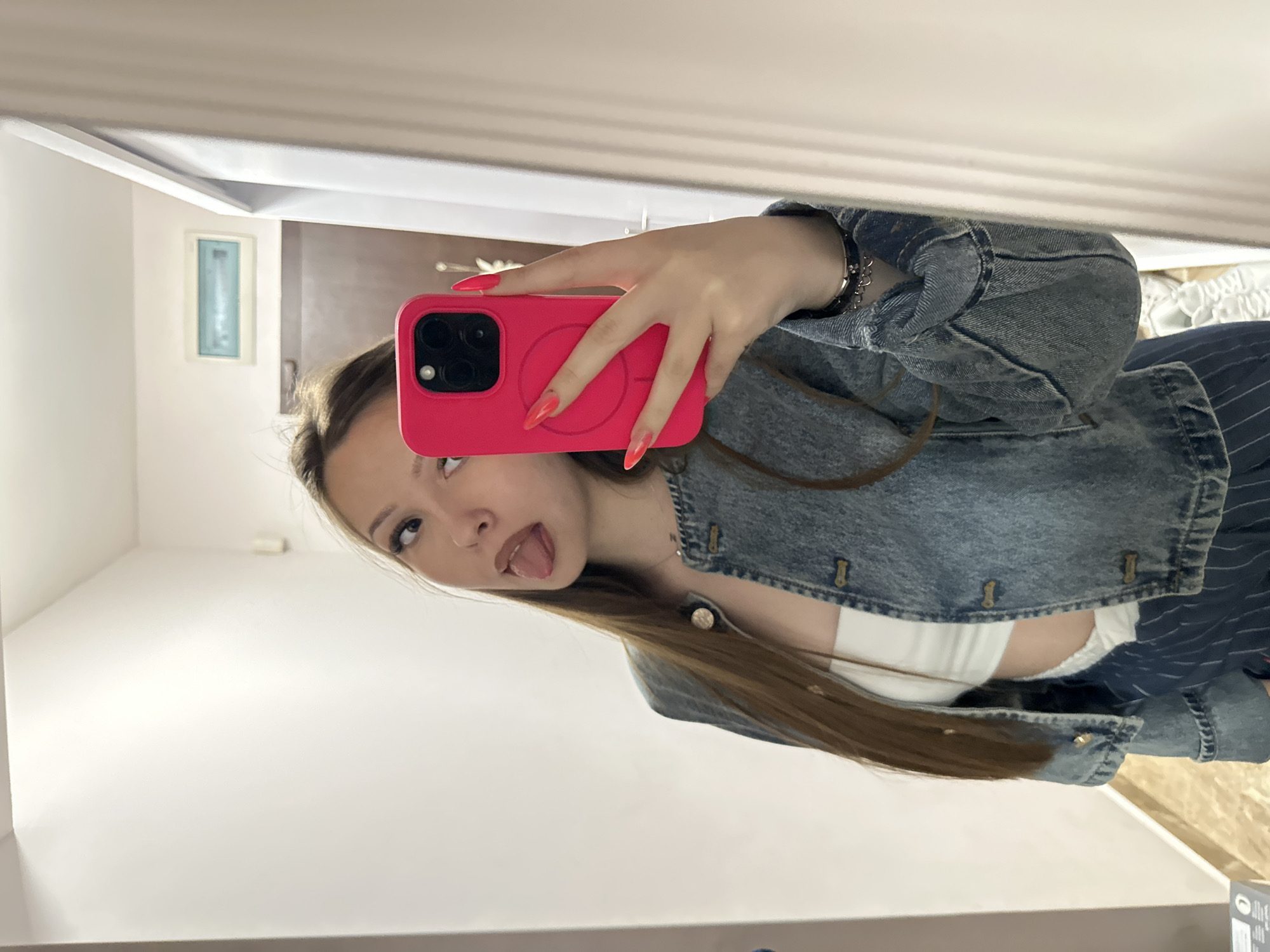 A young woman takes a selfie in front of a mirror. She is wearing a blue denim jacket and a white top underneath. She holds a pink smartphone with long, red fingernails. Her hair is long and brown. She is sticking out her tongue and looking to her left. The reflection shows a white wall with a small, rectangular picture frame on the wall. The background is blurred, but it appears to be a simple interior. There is no visual information to identify the specific location in Varna, Bulgaria.