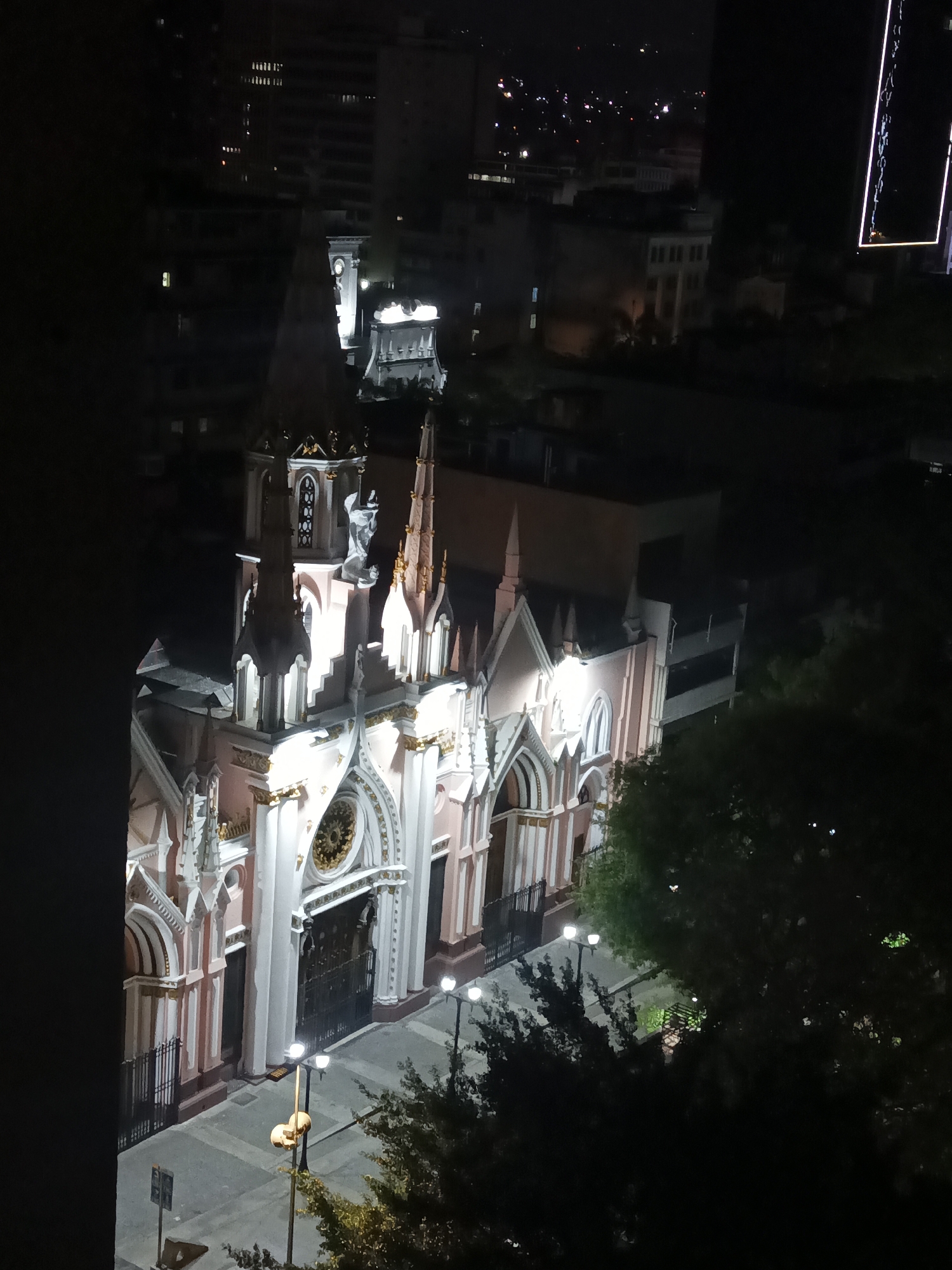The image shows the illuminated Iglesia San Juan Bosco in Caracas, Venezuela, at night. The church has a pink and white facade with golden accents. Its gothic architecture is visible, including spires, arches, and a rose window above the main entrance. The church is surrounded by a paved plaza, with streetlights providing additional illumination. Trees are visible adjacent to the plaza. In the background are urban buildings, some lit, giving context to the church's location within the city.