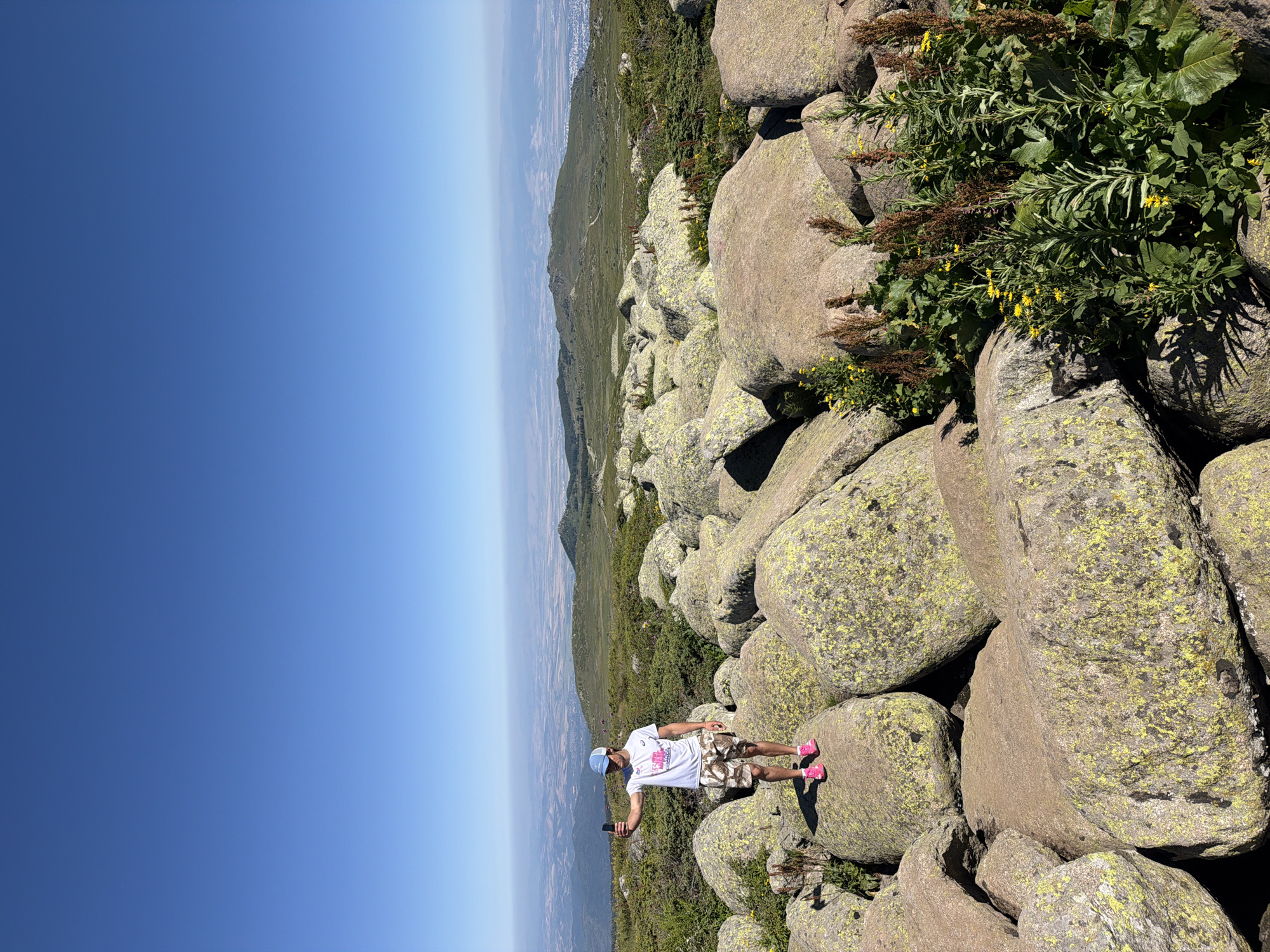 A young man stands on a pile of large, moss-covered boulders, taking a selfie with his phone. He is wearing a white t-shirt, camouflage shorts, a blue baseball cap, and pink athletic shoes. The background reveals a vast, hazy landscape with rolling green hills and a distant city. The sky above is a clear, vibrant blue. The image was taken in the city of Bistritsa, Bulgaria, likely on Vitosha Mountain.