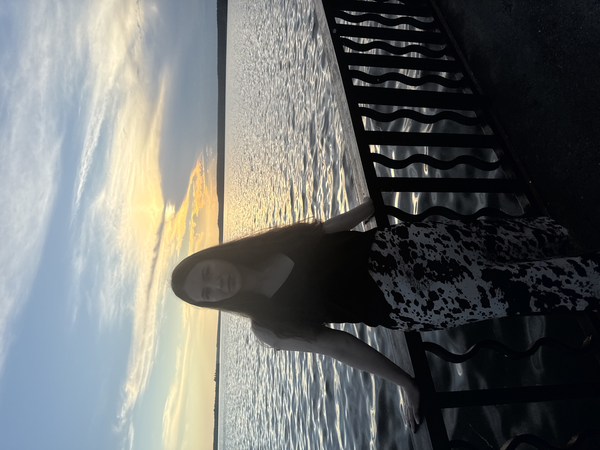 A woman stands on a pier at dusk, leaning on a black metal railing. She has long, dark hair and is wearing a black top and a patterned skirt. The water in front of her is rippling, and the sky is a mix of blue, yellow, and gray clouds, with the sun setting on the horizon. The scene appears to be a serene evening by the water. This location is likely the embankment in Votkinsk, Russia, as the city is known for its picturesque waterfront.