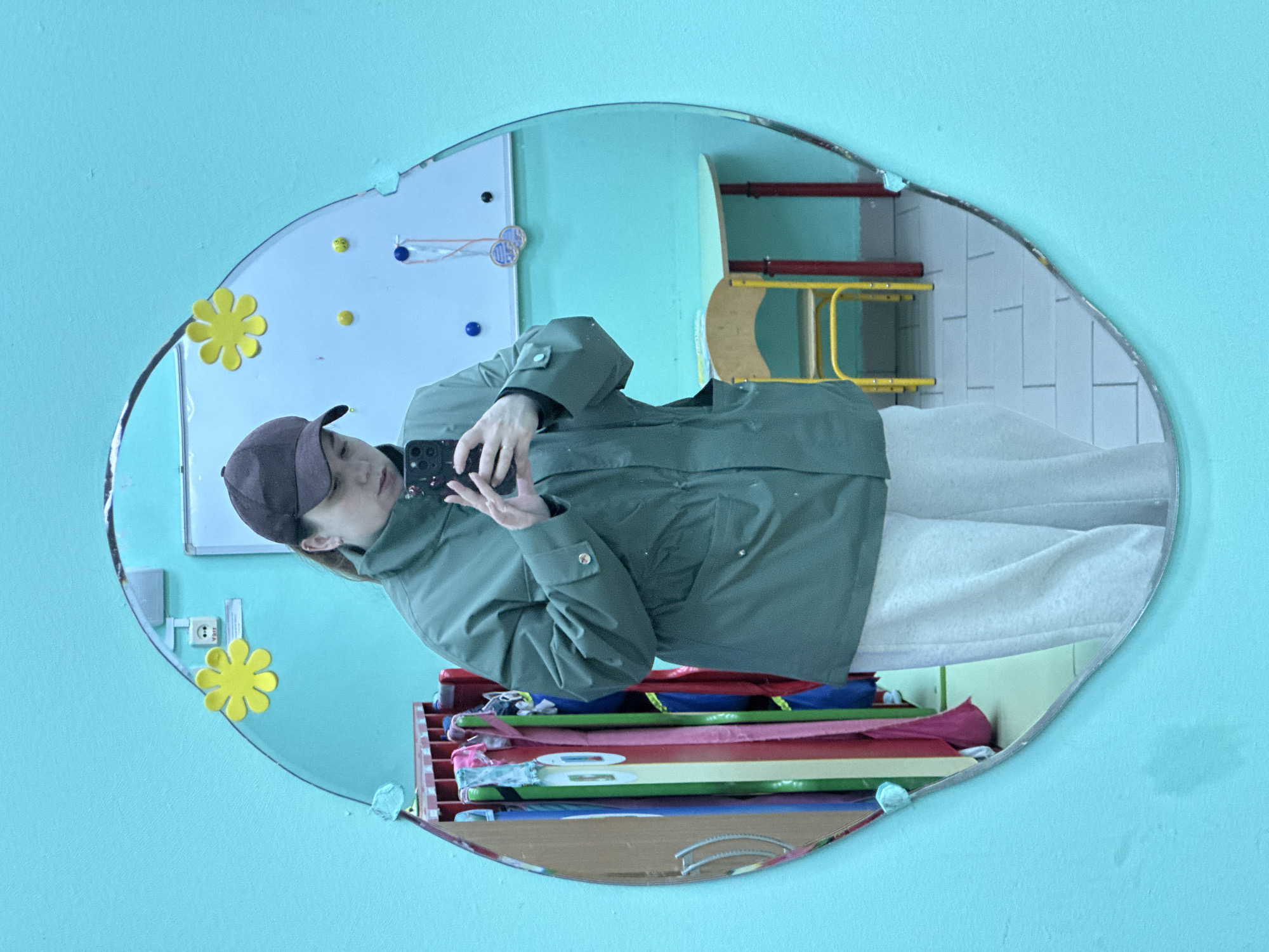 A reflection in a decorative oval mirror mounted on a light blue wall depicts a person holding a smartphone and a cluttered interior space. The person wears a green jacket and a dark cap. The mirror's reflection also shows stacked shelves containing colorful objects and a white board with affixed decorations. A yellow child's chair is visible in the background. The scene suggests a casual, personal photograph taken indoors. The location details are not provided in the image.