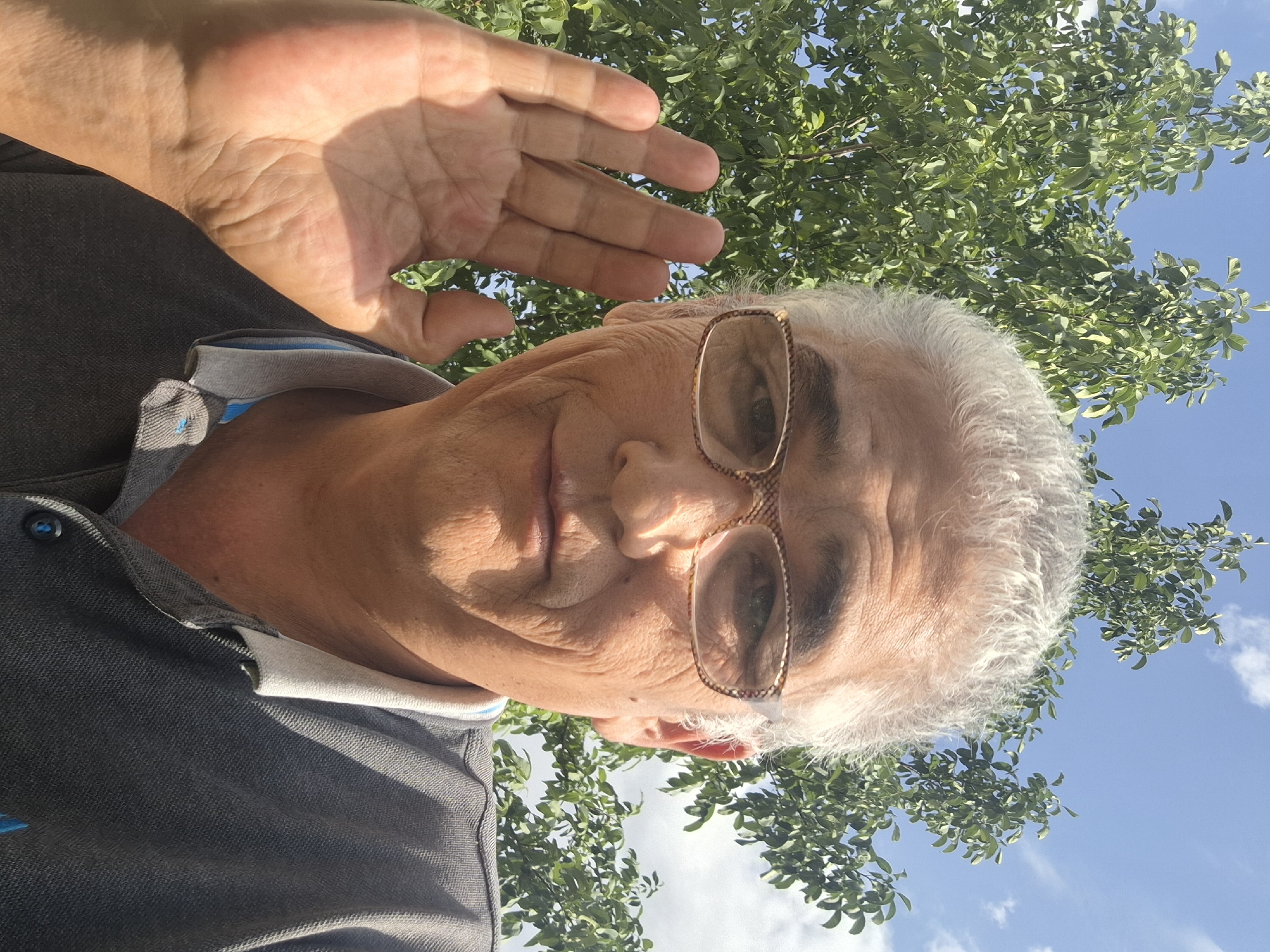 An elderly East Asian man with gray hair and glasses is taking a selfie outdoors. He is wearing a gray polo shirt and has his left hand raised to his ear, as if listening. The background consists of green trees and a blue sky with some clouds. The image was taken in Almaty, Kazakhstan.
