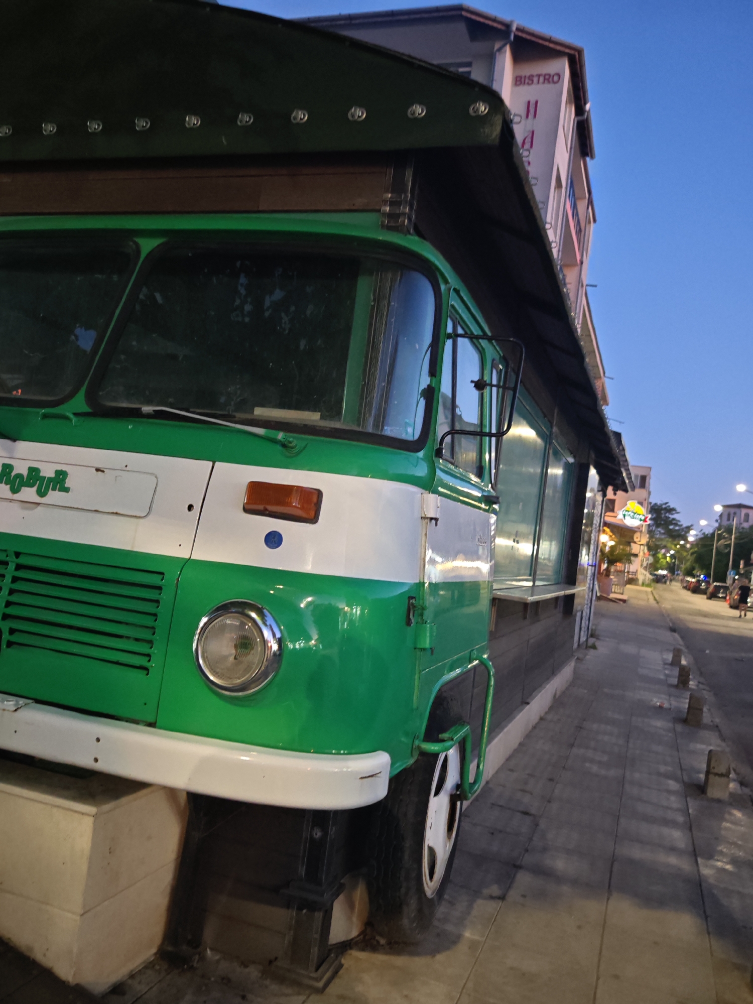 The image depicts a green and white Robur bus-turned-kiosk, likely a stationary food vendor, located on a cobblestone sidewalk in Nesebar, Bulgaria. The bus, with the name "ROBUR" visible, is nestled under a dark, wooden-framed canopy that extends from the side of a building labelled "BISTRO". The bus front is predominantly green, with a white horizontal stripe. The sidewalk runs along a paved street, with sparse pedestrian traffic and parked cars visible in the distance. Streetlights illuminate the street. The sky is a pale blue, indicative of dusk or dawn. The scene has a static, candid quality.