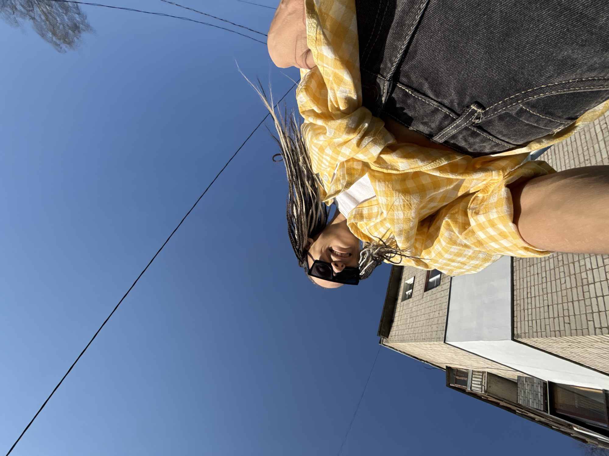 A low-angle shot of a person in Fedorino, Russia, wearing a yellow and white checkered shirt, black denim shorts, and sunglasses. The person has braided hair with some strands dyed white. Behind the person is a clear blue sky with power lines crossing the frame. A building with a brick facade and a white painted section is also visible in the background.