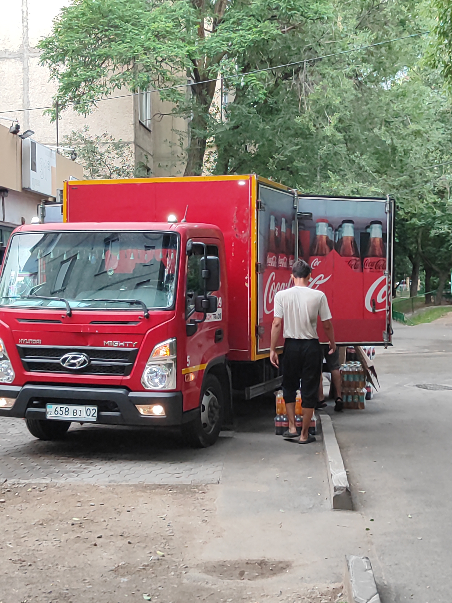 A red Hyundai Mighty truck, likely a delivery vehicle, is parked on a paved surface in an urban setting. The truck's rear doors are open, revealing rows of Coca-Cola bottles stacked in red cardboard cases. A man wearing a white t-shirt and black shorts stands in front of the truck, facing away from the camera, and appears to be interacting with the cargo or an unseen person. To the right of the truck, several cases of bottled drinks are stacked on the ground. The background shows a building with some trees and foliage. The license plate of the truck is visible and reads "KZ 658 BI 02". The scene appears to be outdoors during the daytime, with overcast or shaded lighting. The overall impression is a commercial delivery or distribution event.
