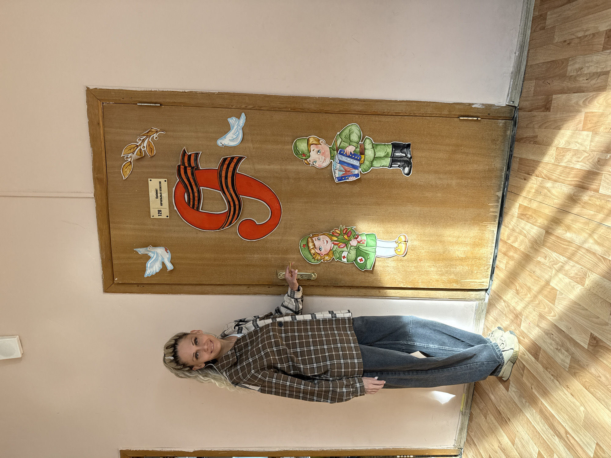 The image shows a woman standing in a hallway. The woman has blonde hair and is wearing a plaid shirt and jeans. She is pointing at a wooden door which is decorated with images of a military theme. The door has an identification tag which reads, "Cabinet 128, Начальная Классов" which is written in Russian. The location is likely in Russia, possibly in Rusanovo, Russia, due to the Cyrillic text and general visual context of the image. The hallway has light-colored walls and wood-patterned linoleum flooring.