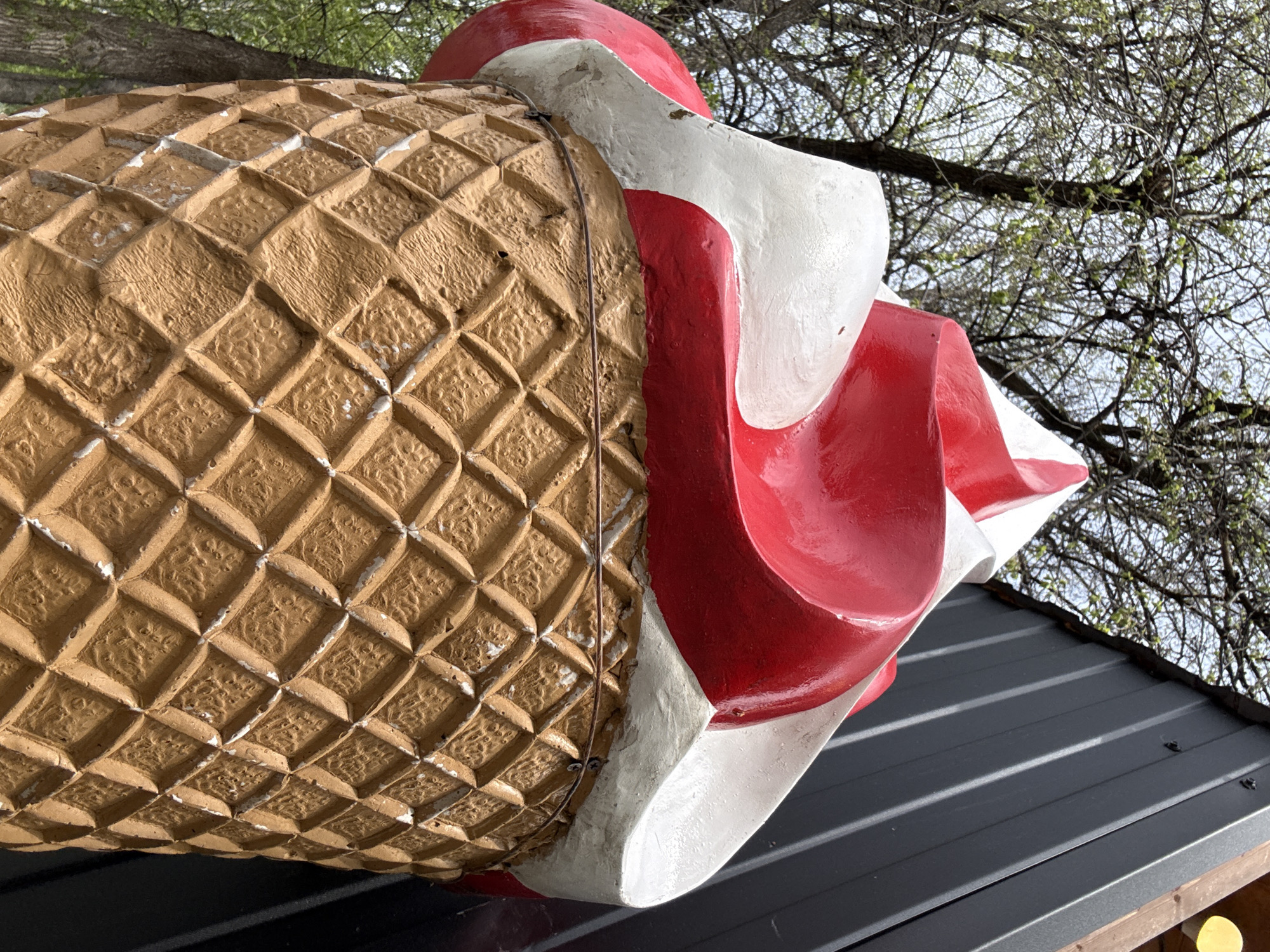 A low-angle, close-up shot depicts a large, decorative ice cream cone mounted on a dark metal roof. The cone is composed of a textured, tan waffle cone section and a red and white swirl of ice cream at the top. A thin wire runs vertically along the side of the cone, possibly used for mounting or support. The roof features a ribbed texture with visible screws. In the background, tree branches with budding leaves are seen against a bright sky. The location is identified as Yekaterinburg, Russia.