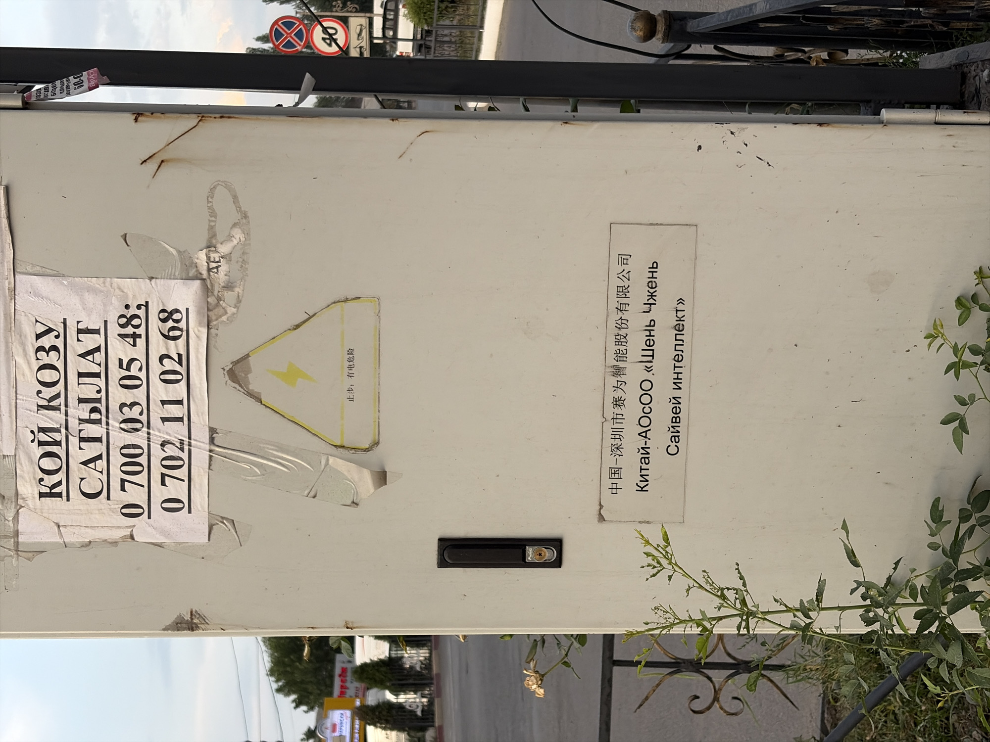 This is a photograph of a metal enclosure with several stickers and labels. The enclosure appears to be outdoors, with a fence and some greenery visible in the background. There are two prominent stickers on the left side of the enclosure. One has text in Cyrillic and appears to be advertising something with phone numbers: "КОЙ КОЗУ САТЫЛАТ 0 700 03 05 48; 0 702 11 02 68". Another sticker is mostly obscured but contains some red and black text.
On the right side of the enclosure, there is a white label with Chinese and Russian text. The Chinese text reads "中国-深圳市赛为智能股份有限公司", which translates to "China-Shenzhen Sifang Smart Co., Ltd.". Below this, the Russian text states "Китай-АОСОО «Шень Чжень Сайвей интеллект»", which appears to be a transliteration and translation of the company name.
There is also a yellow sticker with a black lightning bolt symbol, indicating danger or electricity. Below this symbol, in Chinese characters, it says "止步:有电危险", meaning "Stop: danger of electricity". A black lock mechanism is also visible on the enclosure.
In the background, there are traffic signs, including a circular sign with a red border and a diagonal red bar, and another sign with the number "40" inside a circle. The overall scene suggests an industrial or utility area, possibly near a road. The lighting suggests it is daytime. The presence of multiple languages on the labels indicates an international context or a place with diverse linguistic influences.
