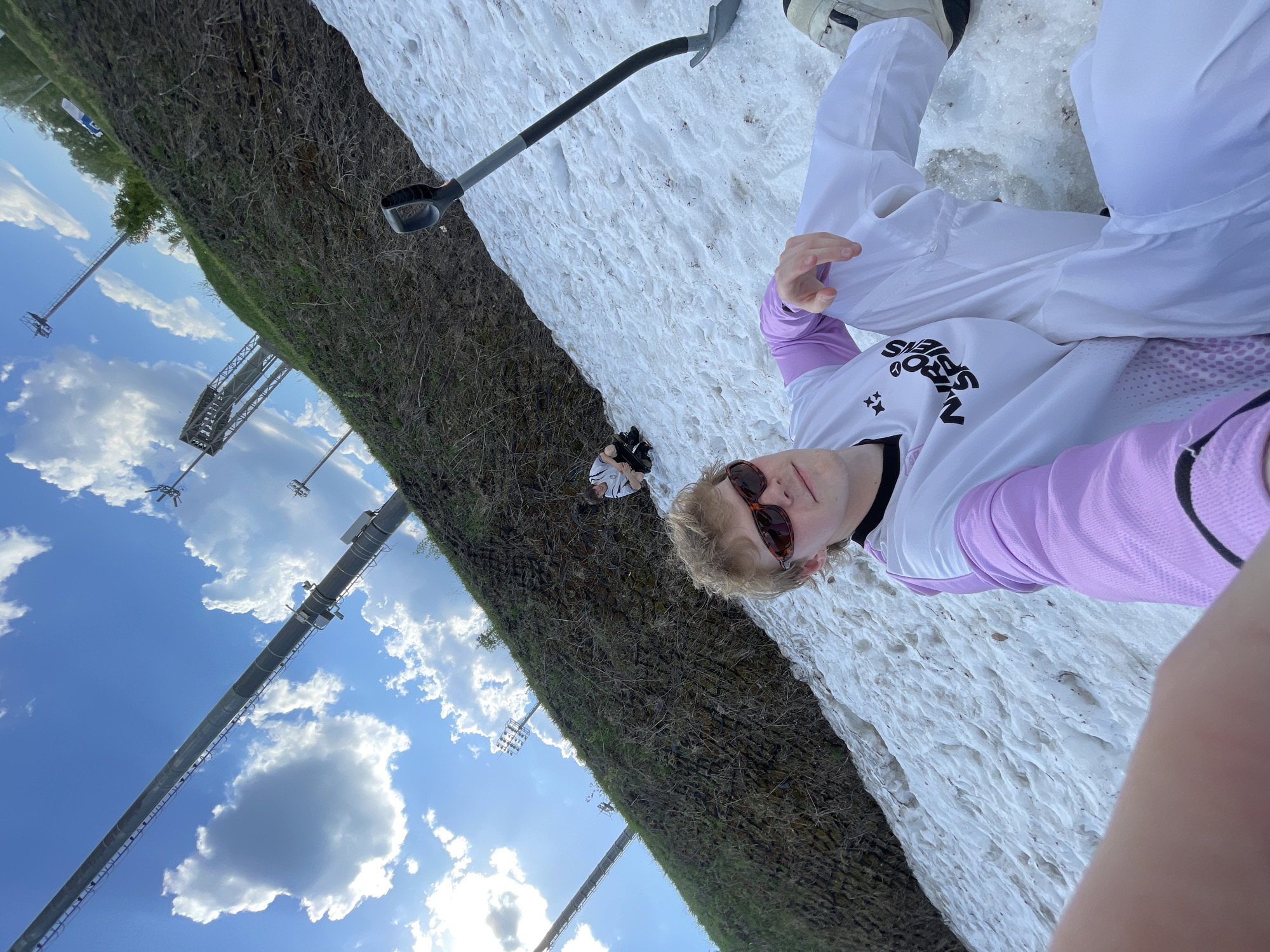 A young person in sunglasses, a white and light purple long-sleeved shirt, and white pants, sits on a snow patch next to a grassy area. A shovel rests on the snow. Another person is partially visible in the distance on the border between the snow and grass. The background includes stadium lighting and a blue sky with white clouds. The image appears to be taken in Krasnoyarsk, Russia, given the location metadata.