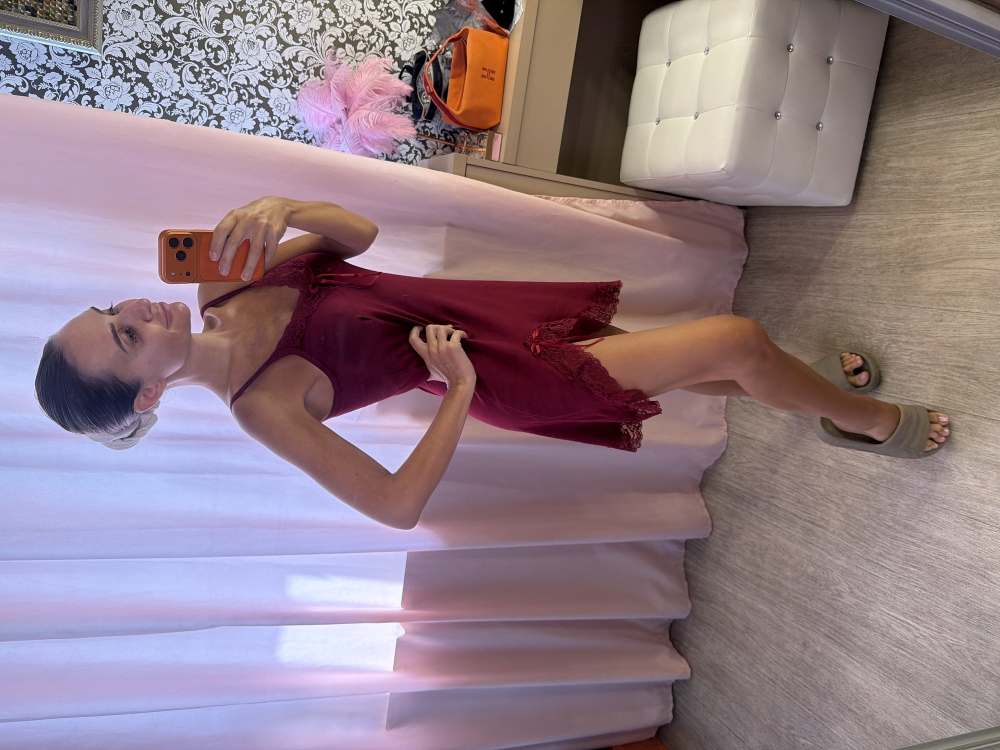A woman stands in a room, taking a mirror selfie with her phone. She is wearing a short, dark red slip dress with lace trim and bows. She has her hair pulled up in a bun and is wearing grey slide sandals. The room has light grey wood-look flooring and is decorated with pink curtains on the left and patterned wallpaper on the right. A white ottoman with rhinestone accents is visible to the right of the woman. A dresser with an orange handbag and a pink feather boa is also present. The overall impression is a bedroom or dressing area.
