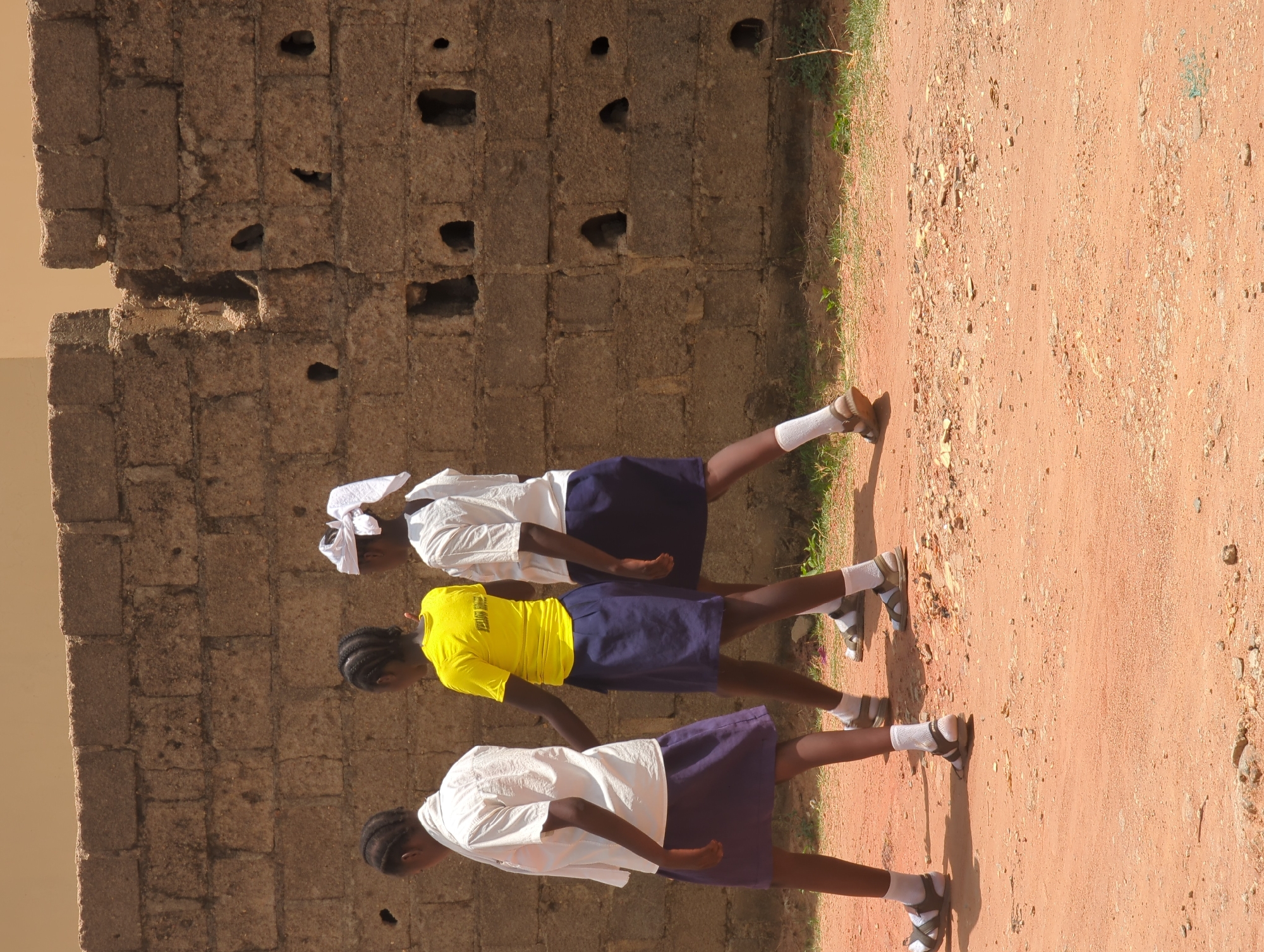 Three girls wearing school uniforms walk along a dirt path next to a weathered brick wall. The girl closest to the camera is wearing a white shirt and a blue skirt. The middle girl is wearing a yellow shirt and a blue skirt. The girl furthest from the camera is wearing a white shirt and a blue skirt, with a white head covering. All three girls are wearing white socks and sandals. The scene is located in Narayi, Nigeria.