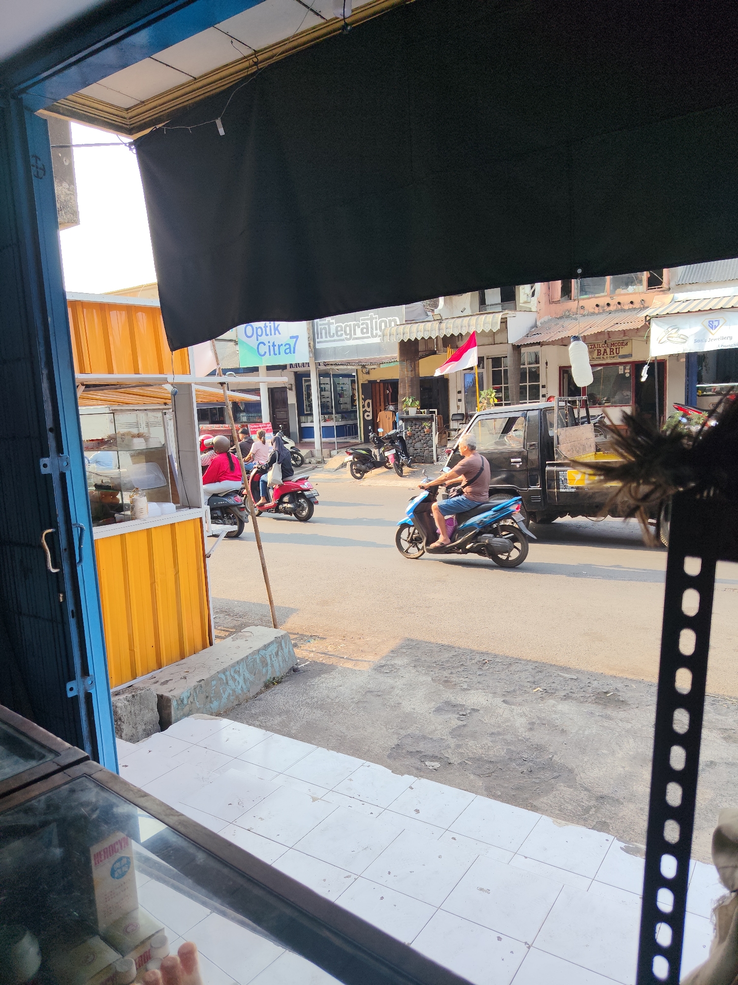 In Dampit, Indonesia, a street scene unfolds with a focus on vehicular activity and storefronts. A man on a blue motorcycle rides past a parked black truck, while another motorcycle, red, carries two individuals further down the street. The roadside features a yellow food stall, accompanied by a grey concrete curb and a paved walkway. The buildings lining the street host various businesses, including "Optik Citra7" and "TAILOR MODE BARU," with awnings providing shade over the shop fronts. The Indonesian flag is visible, fluttering near one of the establishments. The lighting suggests it is daytime, casting shadows on the ground. The foreground shows a glimpse of items inside a glass-fronted cabinet.