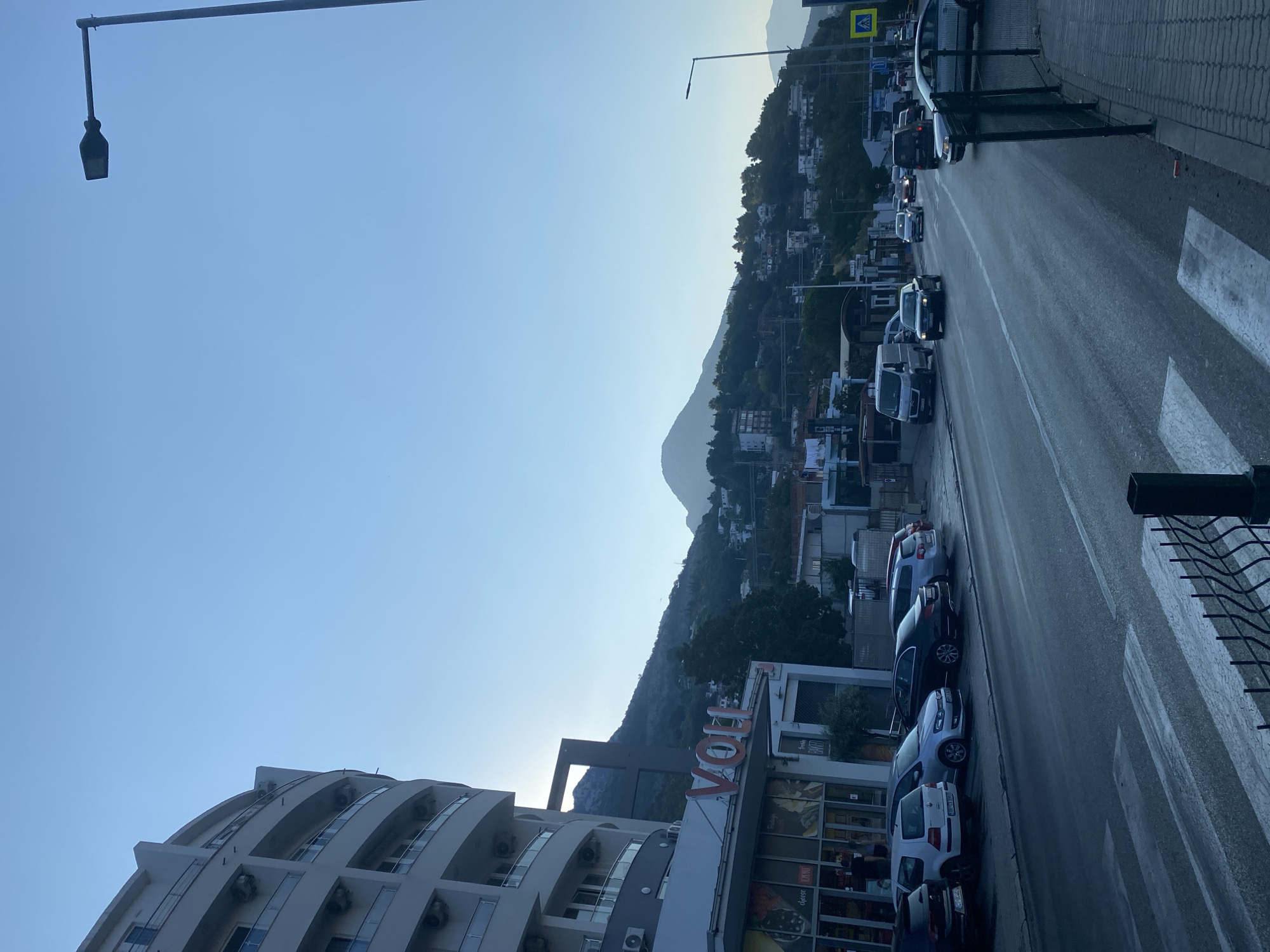 The image captures a street scene in Sutomore, Montenegro, during what appears to be late afternoon or early evening. The foreground features a pedestrian crossing on a wet asphalt road, with a black metal fence to the right. Several cars are parked along the sidewalk, including a white Volkswagen Golf and a silver Volkswagen Polo. Adjacent to the parked cars is a modern, multi-story building with a curved facade. The ground floor of this building houses a store with large glass windows, displaying food items and bearing the signage "VOLI".
Across the street, traffic moves along a multi-lane road. A dark-colored van and several cars, including a black Mercedes-Benz sedan, are visible. In the background, a hillside settlement is dotted with buildings and surrounded by lush green vegetation. The backdrop is dominated by rolling mountains under a clear, pale blue sky. A street lamp is positioned on the right side of the frame, and a yellow pedestrian crossing sign is visible further down the road. The overall lighting suggests the sun is low on the horizon, casting a soft glow over the scene. No people are clearly identifiable or engaged in any specific actions.