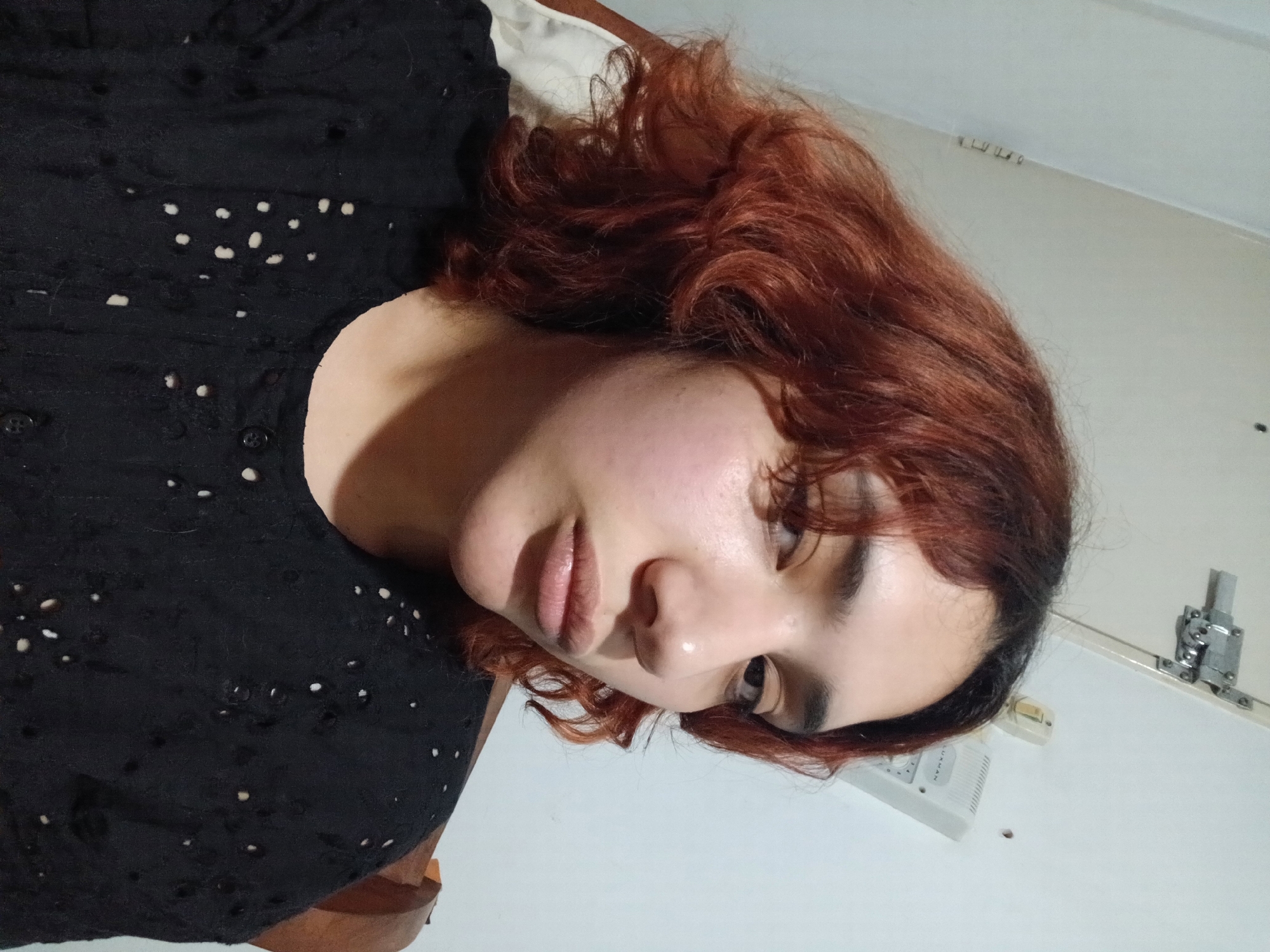 A young woman with reddish-brown curly hair and fair skin is lying down, wearing a black shirt with small cut-out details. She is looking slightly downward with her eyes open. The background is a plain white wall with a white rectangular object (possibly an electrical outlet or a part of a ventilation system) and a metal fixture. The location is indoors. The city of Buenos Aires, Argentina cannot be determined from the provided image.