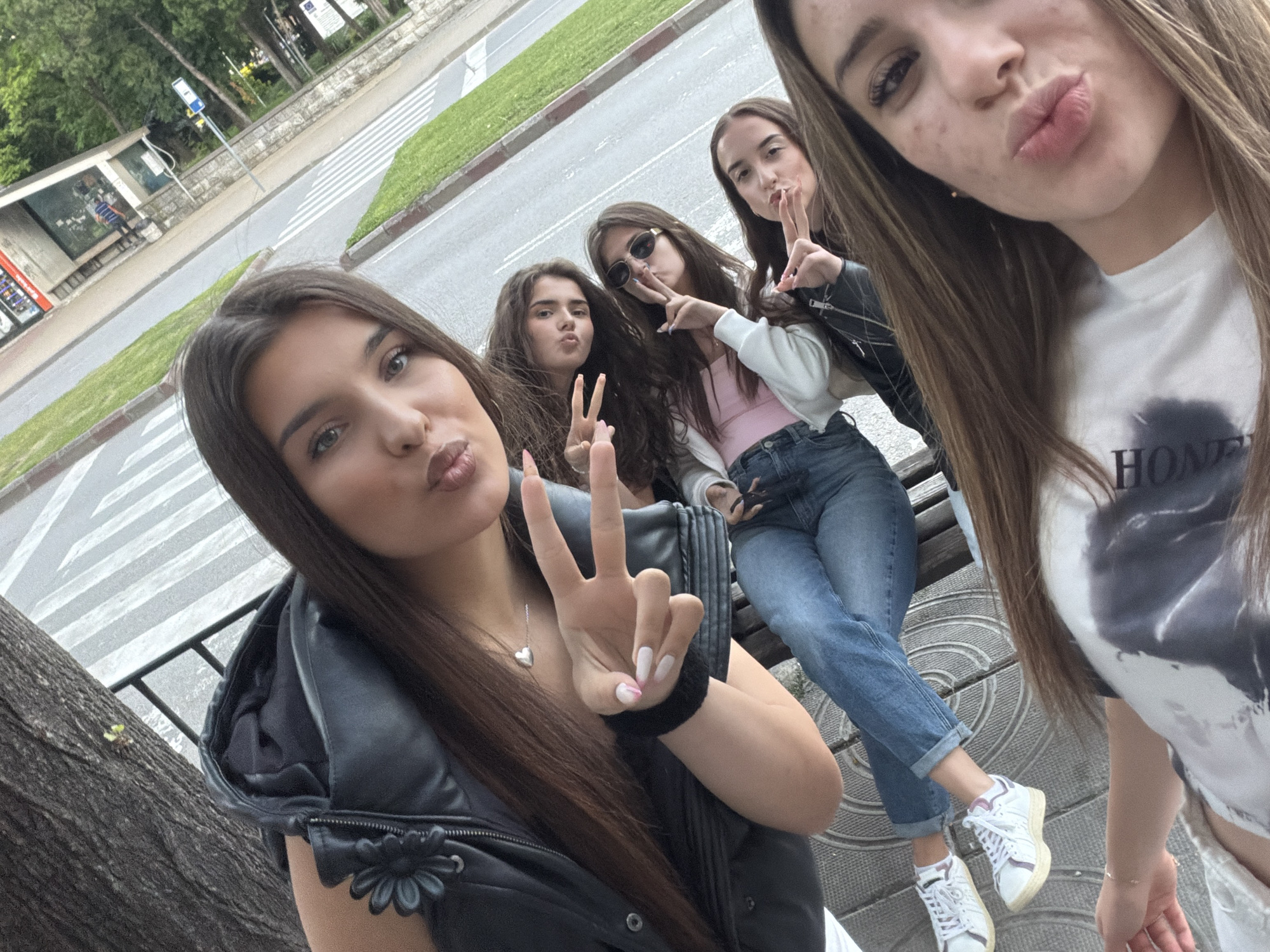 In an outdoor, urban setting, five young women are captured in a selfie on a bench. The foreground is dominated by the face of the woman taking the picture, who has long, light brown hair and is wearing a white t-shirt with a grey and black graphic design. To her left, a second woman, also with long light brown hair, is visible from the chest up, wearing a black leather jacket with a floral embellishment. She is holding up two fingers in a peace sign. Behind her, a third woman with dark brown hair is wearing sunglasses and a pink top, also making a peace sign. Next to her, a fourth woman with long dark brown hair, wearing a black leather jacket, is partially visible. In the background, a fifth woman is mostly obscured by the others. The women are seated on a patterned bench. Behind them, a street with a pedestrian crossing is visible, lined with trees and a building structure, possibly a bus stop shelter. The lighting suggests it is daytime.